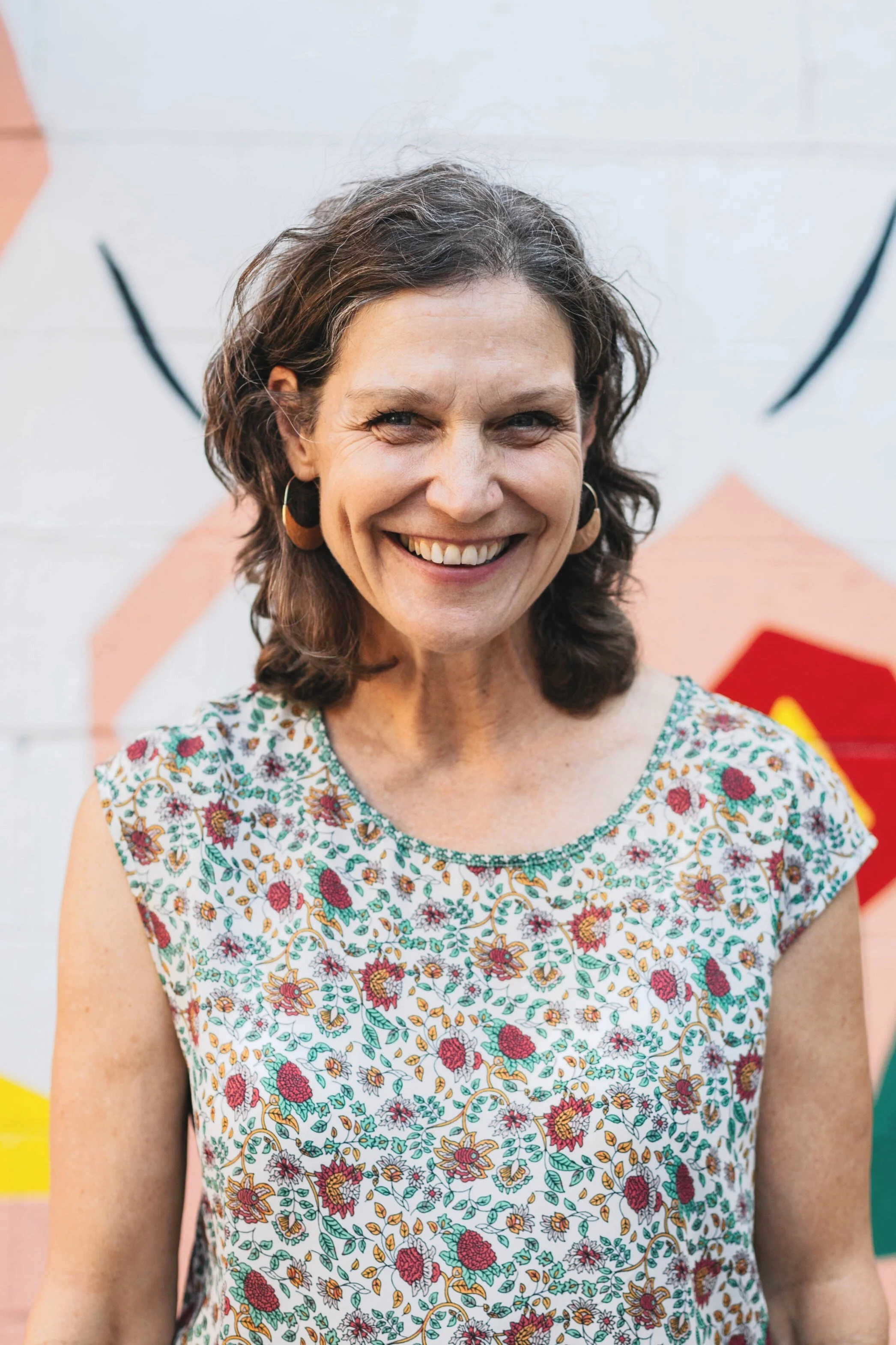 Smiling woman with shoulder-length curly hair, wearing hoop earrings and a sleeveless, floral-printed top, standing in front of a wall with colorful abstract artwork.