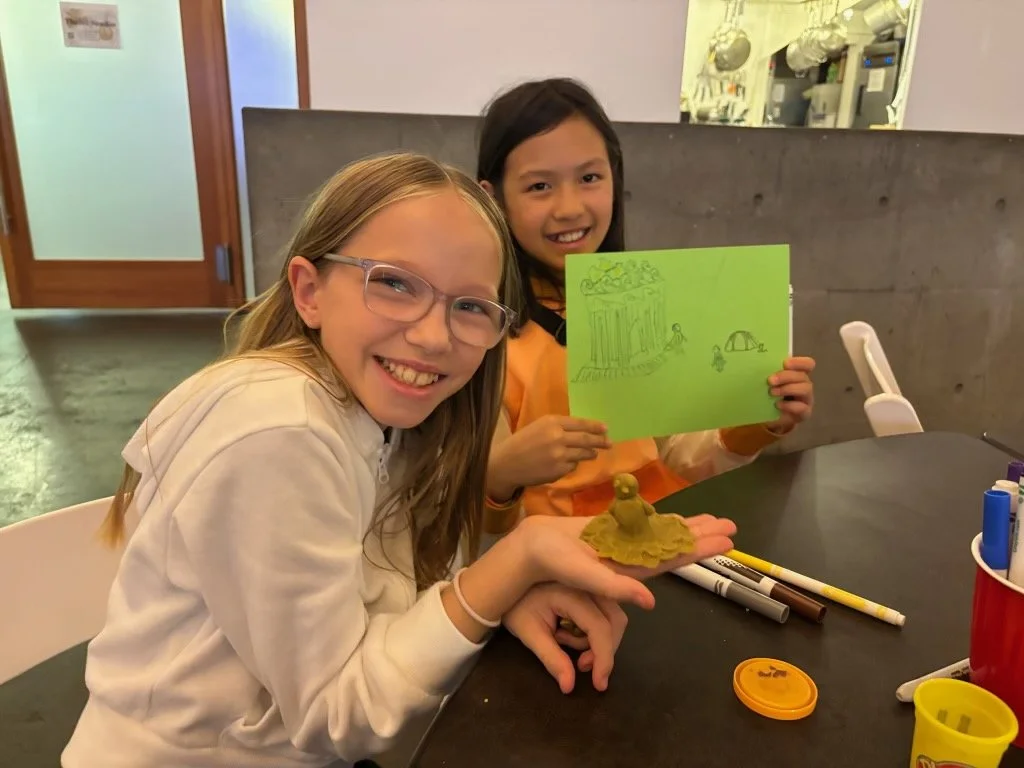 Two young girls are sitting at a table, smiling at the camera. The girl on the left is holding a small yellow chick figurine, and the girl on the right is holding a green drawing or card with a tree, a person, and a car sketched on it.