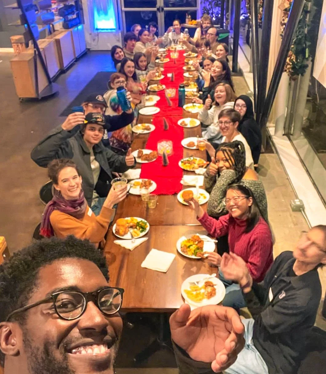 The Union youth group is all gathered around a long banquet table that has a red runner down the middle and everyone has plates of food. Everyone is looking at the camera and smiling as a man holds the camera and takes a selfie.