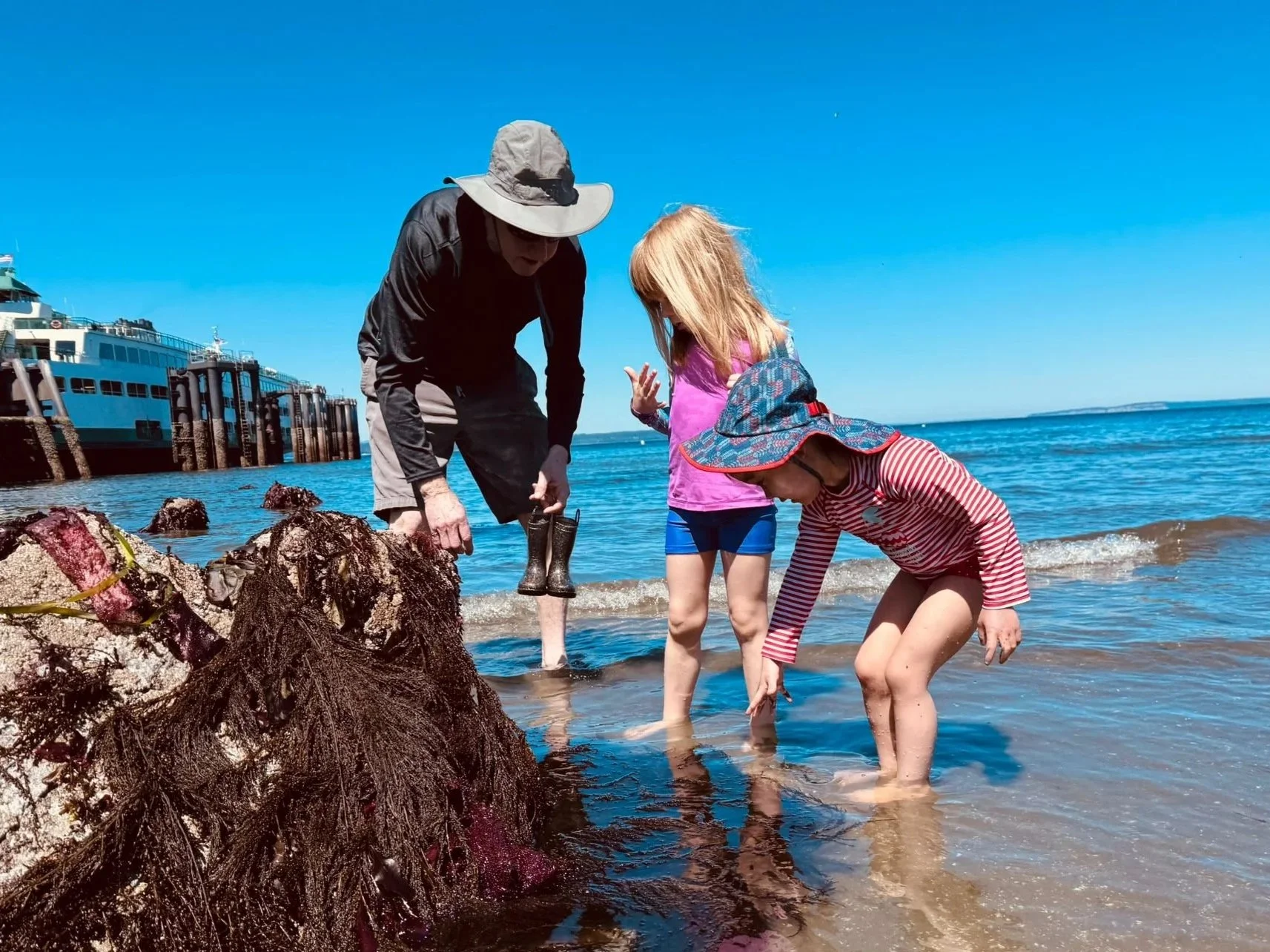 A man and two young children crouch together at a rocky beach tidepool during a Play & Learn summer outing, leaning in with curiosity to explore their discovery