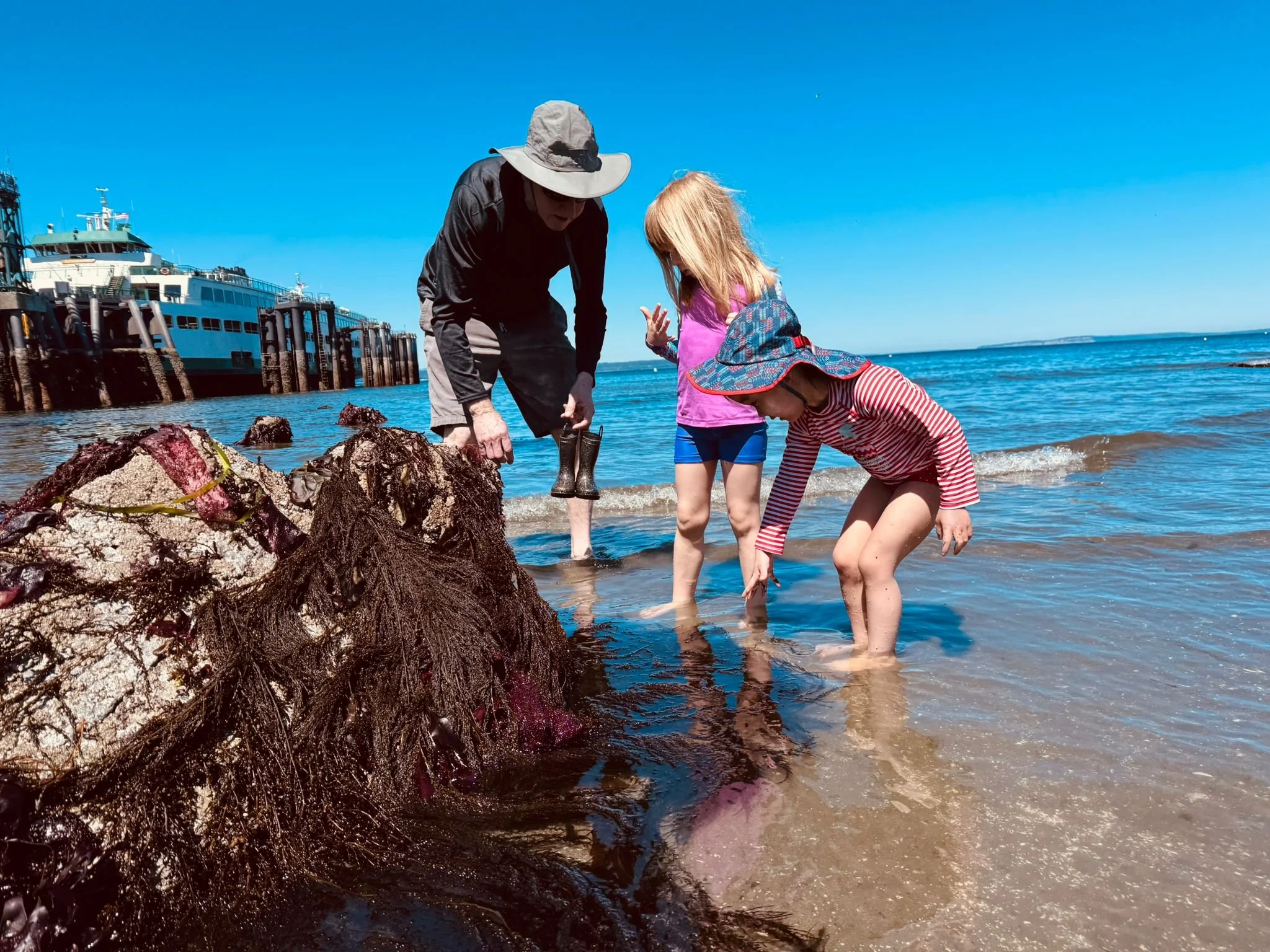 A man and two young children crouch together at a rocky beach tidepool during a Play & Learn summer outing, leaning in with curiosity to explore what lives inside