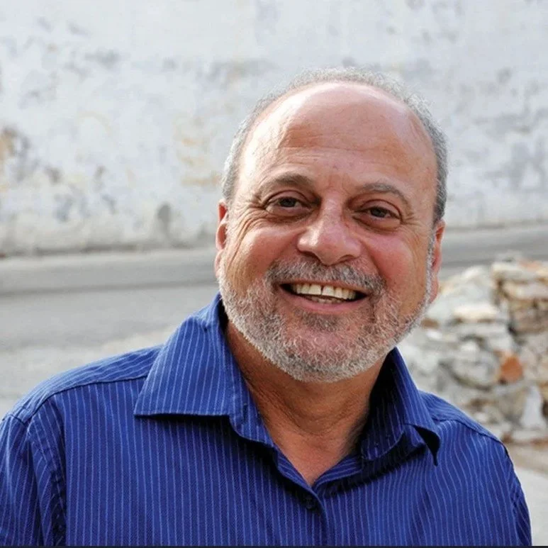 A portrait of a Palestinian man with a close shave beard, and blue button up shirt, smiling with a concrete wall blurred in the background. He is the founder of a reconciliation organization in Palestine and Israel.