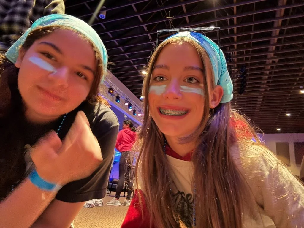 Two girls with blue under eye make smeared on their cheeks, smiling at the camera. Wearing bandana's and beaded necklaces.