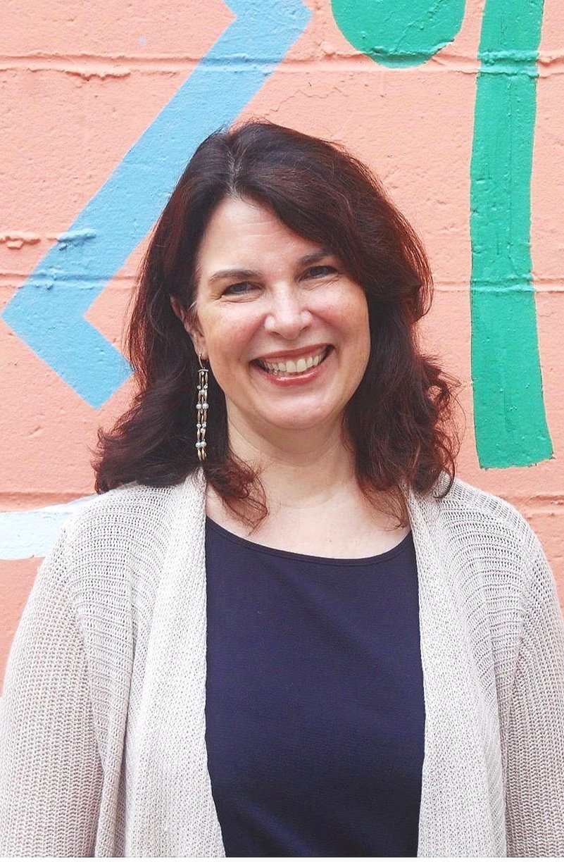 A woman smiling in front of a colorful painted brick wall.