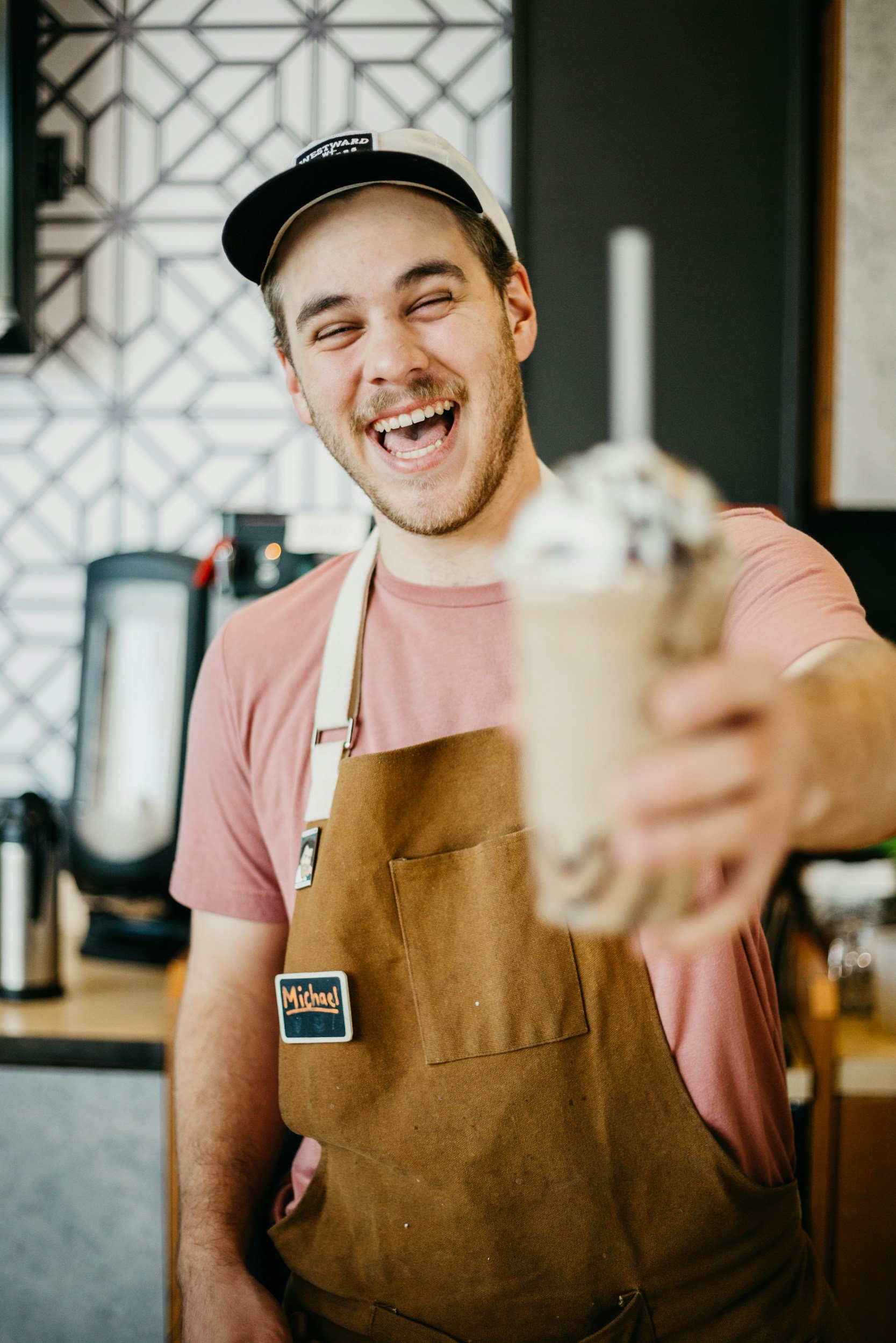 A smiling man wearing a brown apron with a name tag 'Michael' presenting a milkshake topped with whipped cream and chocolate, in a coffee shop or cafe setting