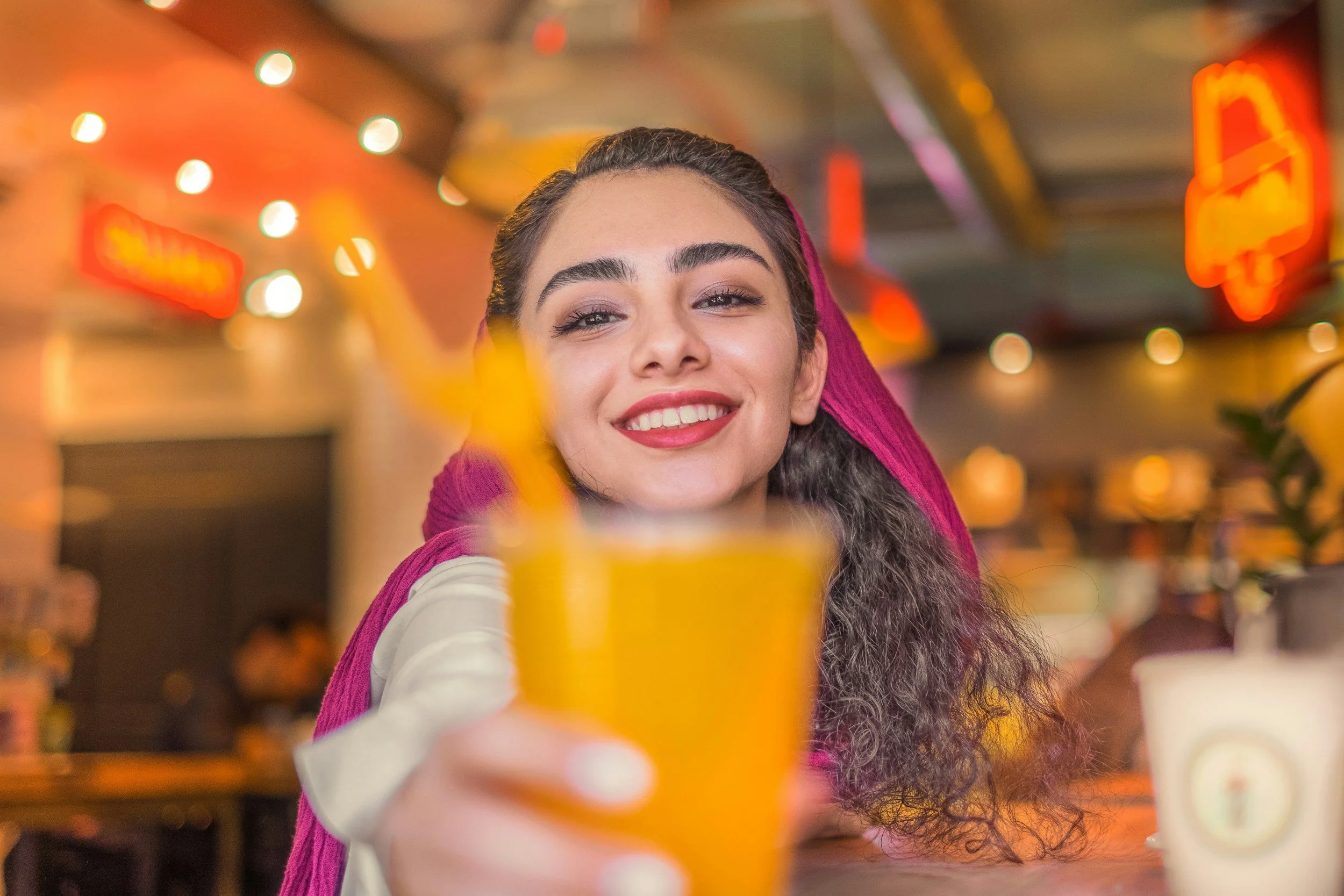 A young woman with long dark curly hair and a pink headscarf smiling and holding a yellow drink with a striped straw at a lively indoor cafe or bar. The background features warm lighting and blurry patrons.