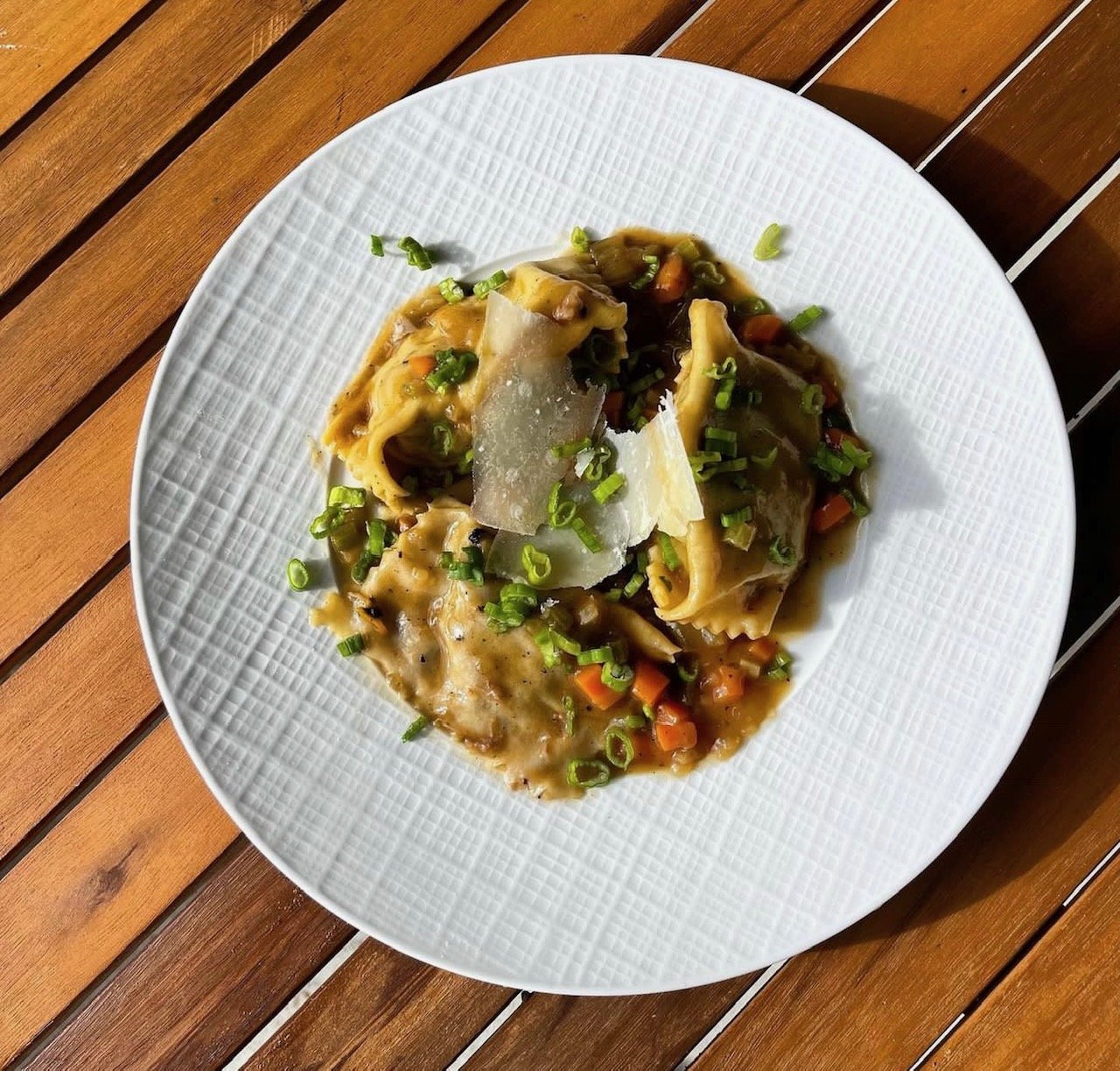 A white textured plate with cooked pasta, vegetables, and shaved cheese, garnished with chopped green onions on a wooden table.