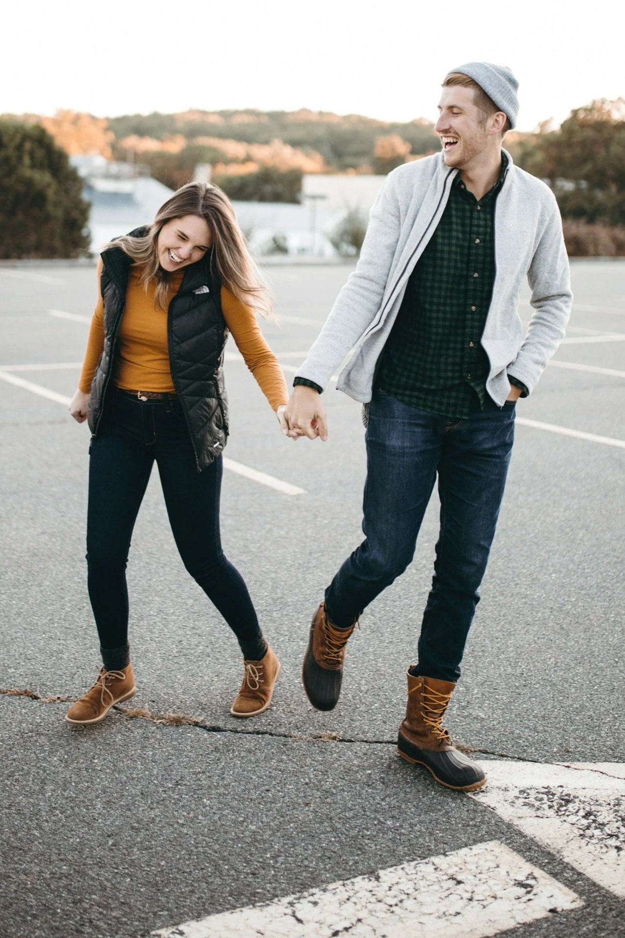 A young man and woman holding hands and walking through a parking lot, both smiling and laughing.