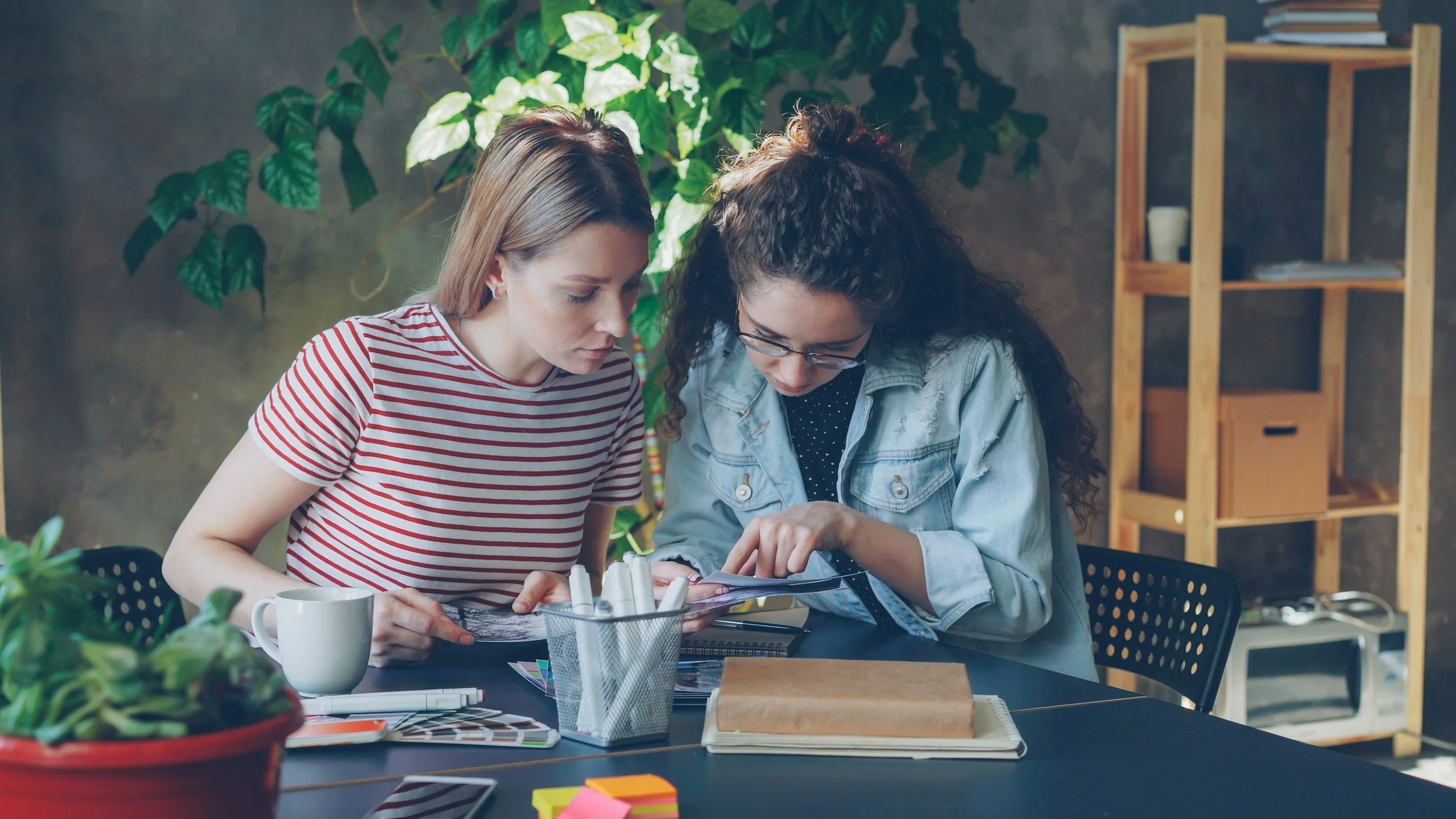 Two women working together at a desk, looking at a tablet, surrounded by design and office supplies, with a plant and shelves in the background.