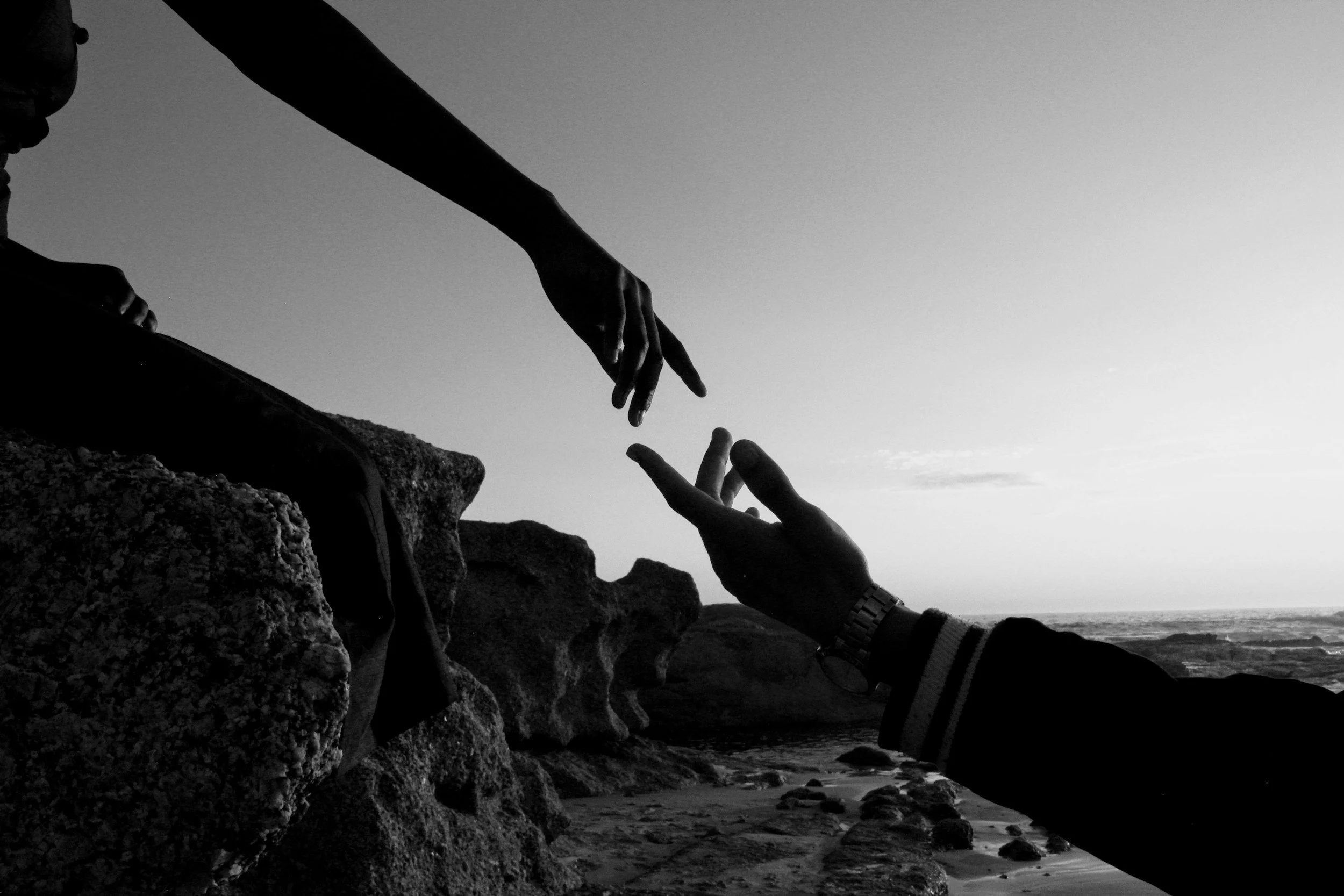 Silhouettes of two hands reaching out towards each other over rocks, with the ocean and sky in the background, black and white photograph.