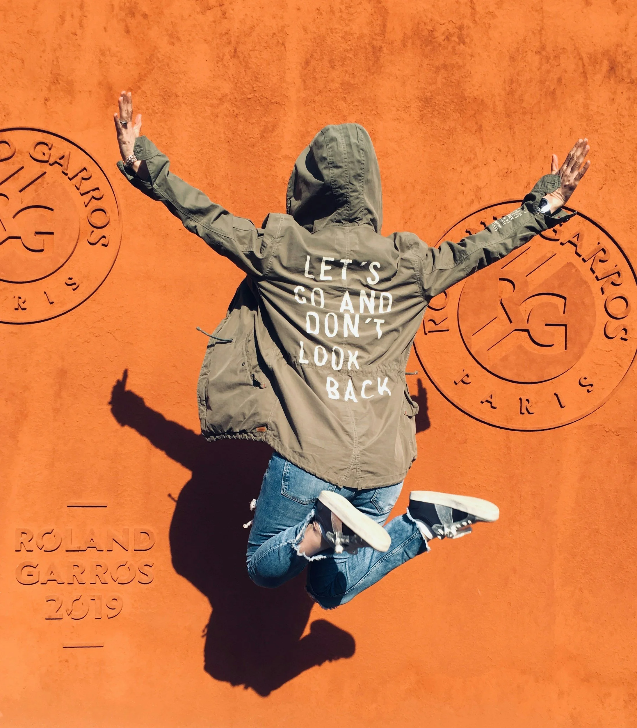 Person jumping in the air with arms outstretched against an orange wall with 'Roland Garros Paris 2019' logo and text, wearing a green jacket with 'Let's go and don't look back' written on the back.