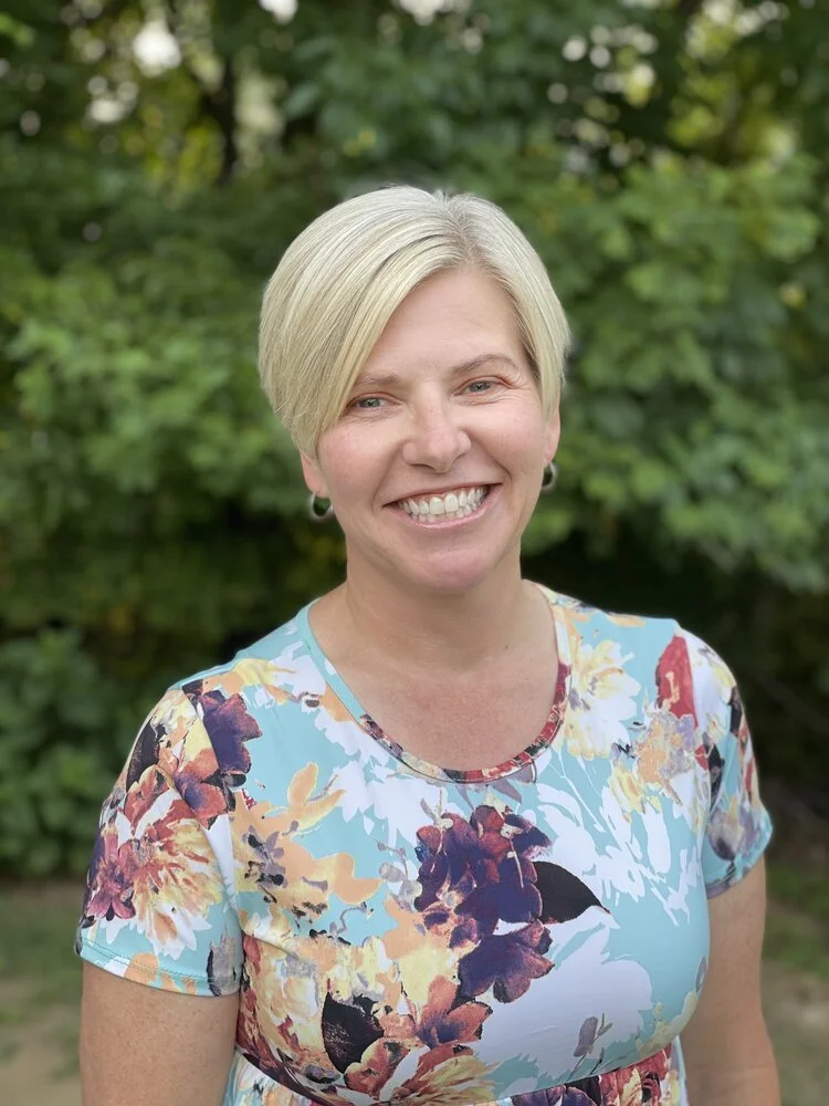 A smiling woman with short blonde hair wearing a floral dress standing outdoors with green trees in the background.