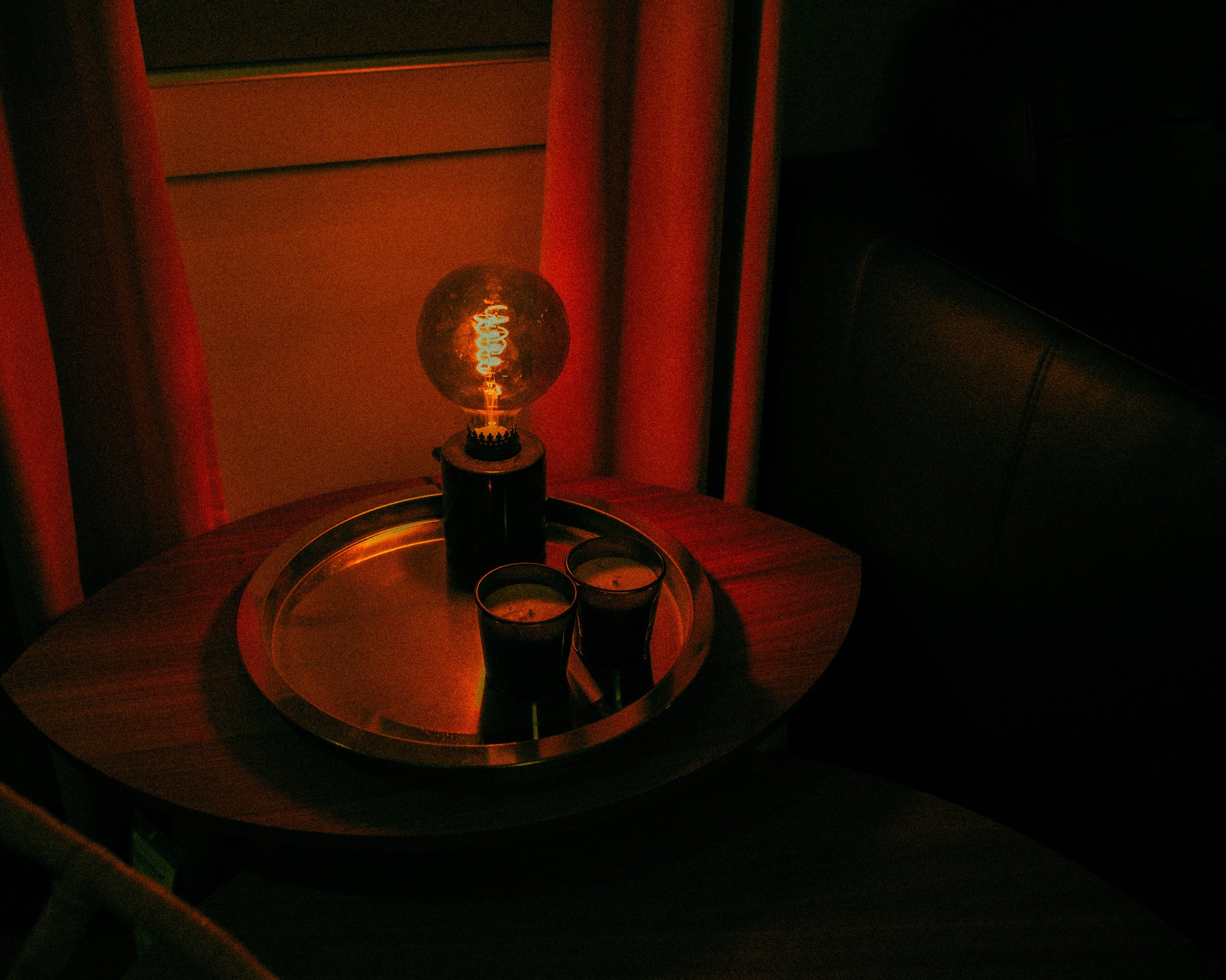 A nightstand with a glowing vintage-style Edison bulb lamp and two glasses of dark beverage on a silver tray in a dimly lit room.