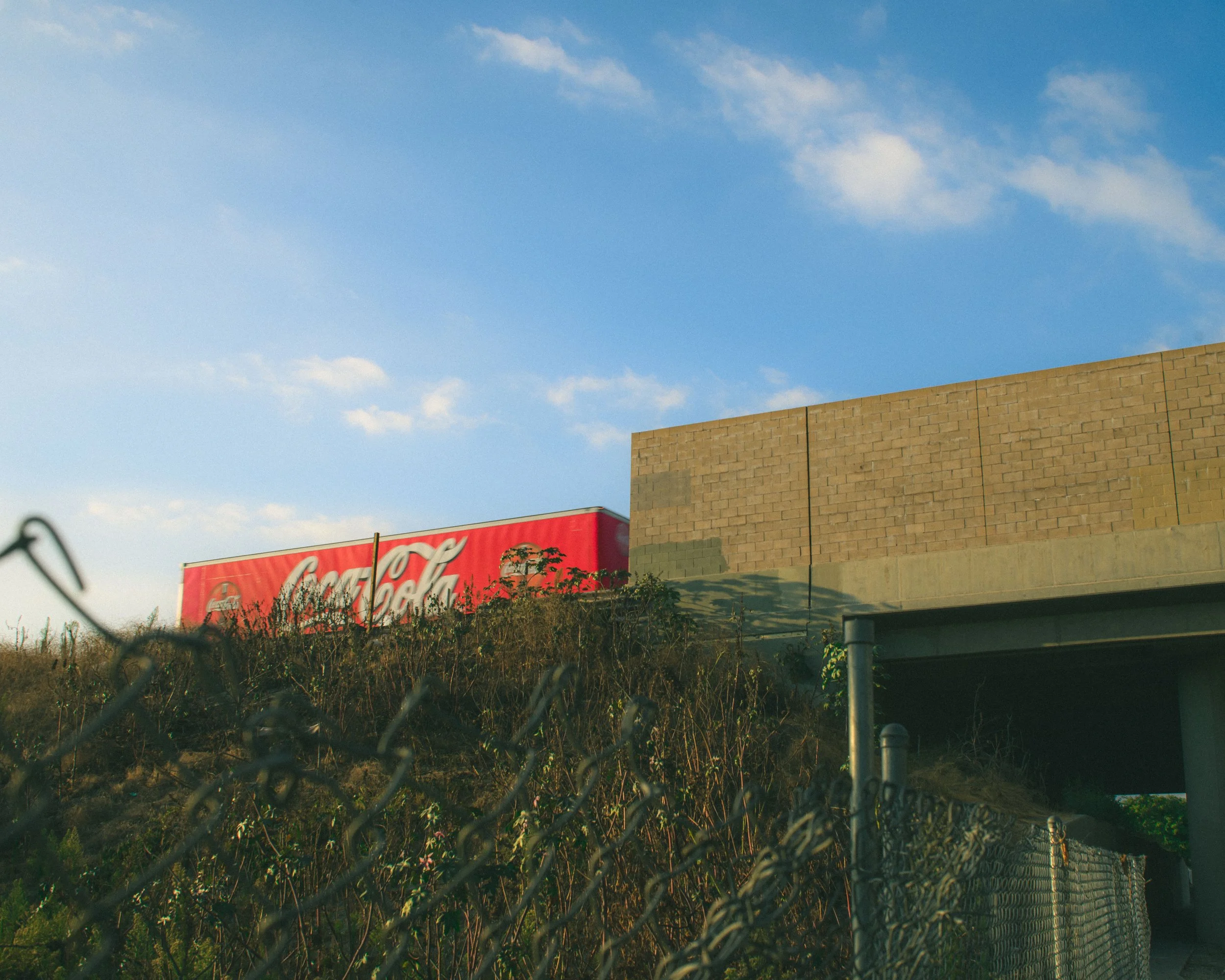 A red Coca-Cola billboard on top of a partially covered building behind a chain-link fence with overgrown bushes, under a blue sky with scattered clouds.