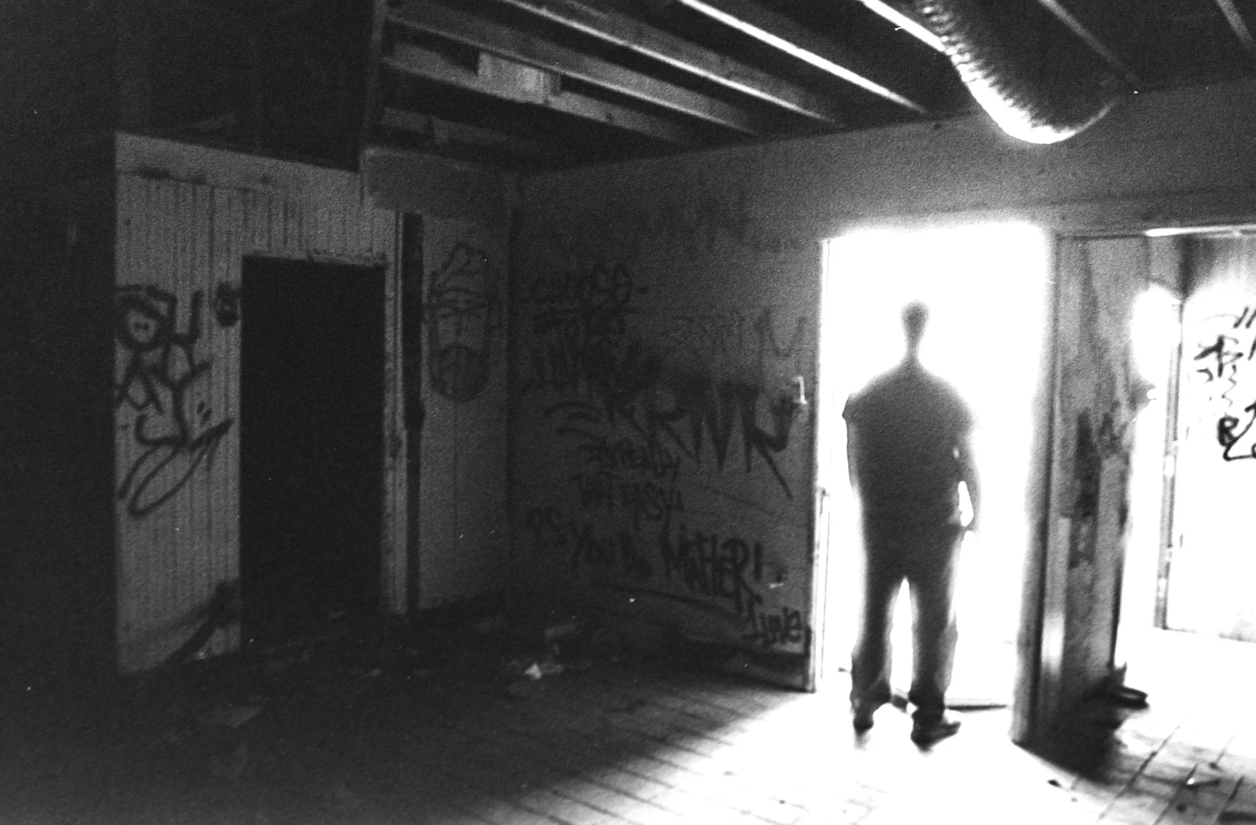 Black and white photo of an interior space with graffiti on the walls, a doorway, and a person standing in the doorway illuminated by bright light from outside.