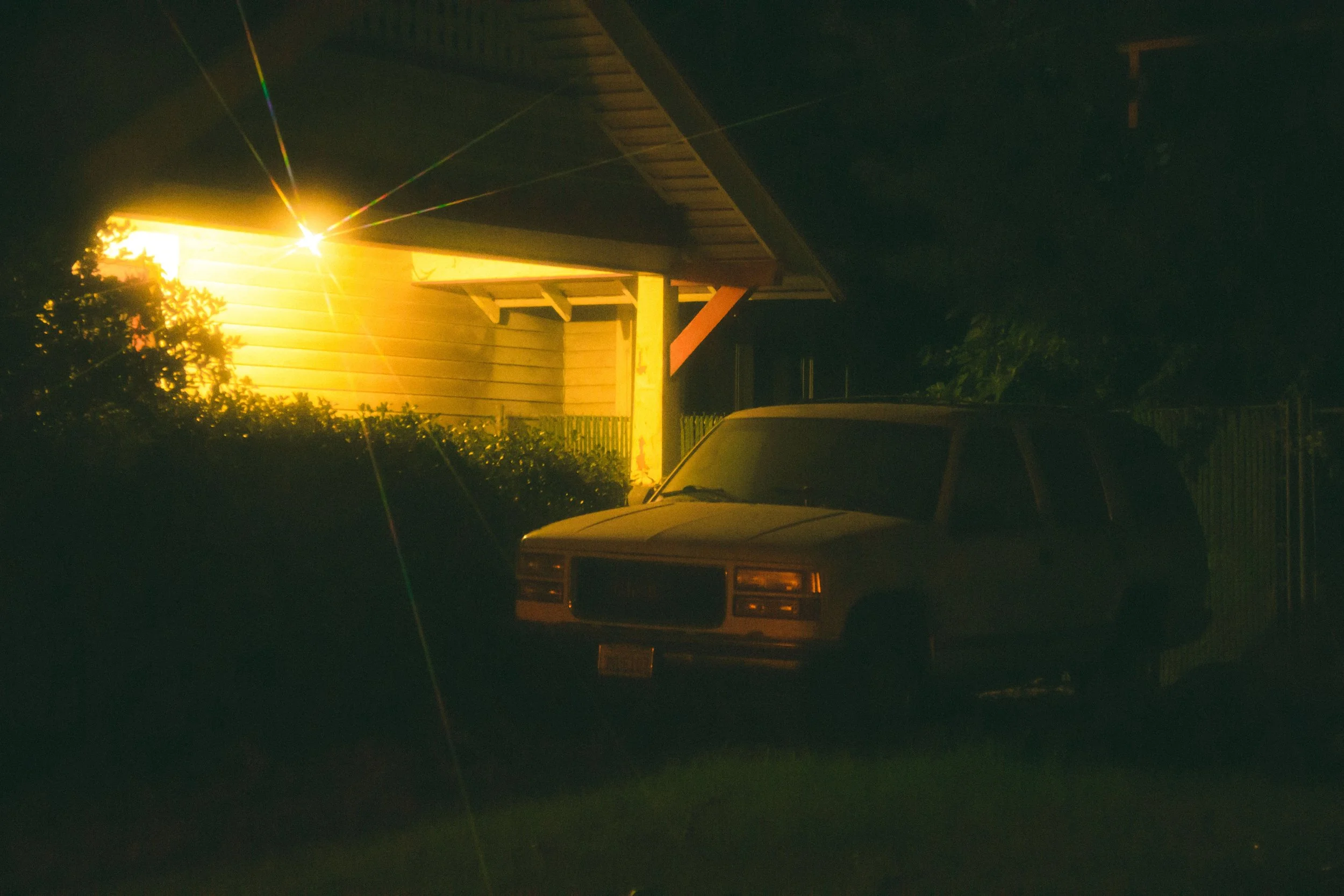A dark night scene showing a parked old SUV under a porch, illuminated by a warm exterior light from a house, with greenery nearby.