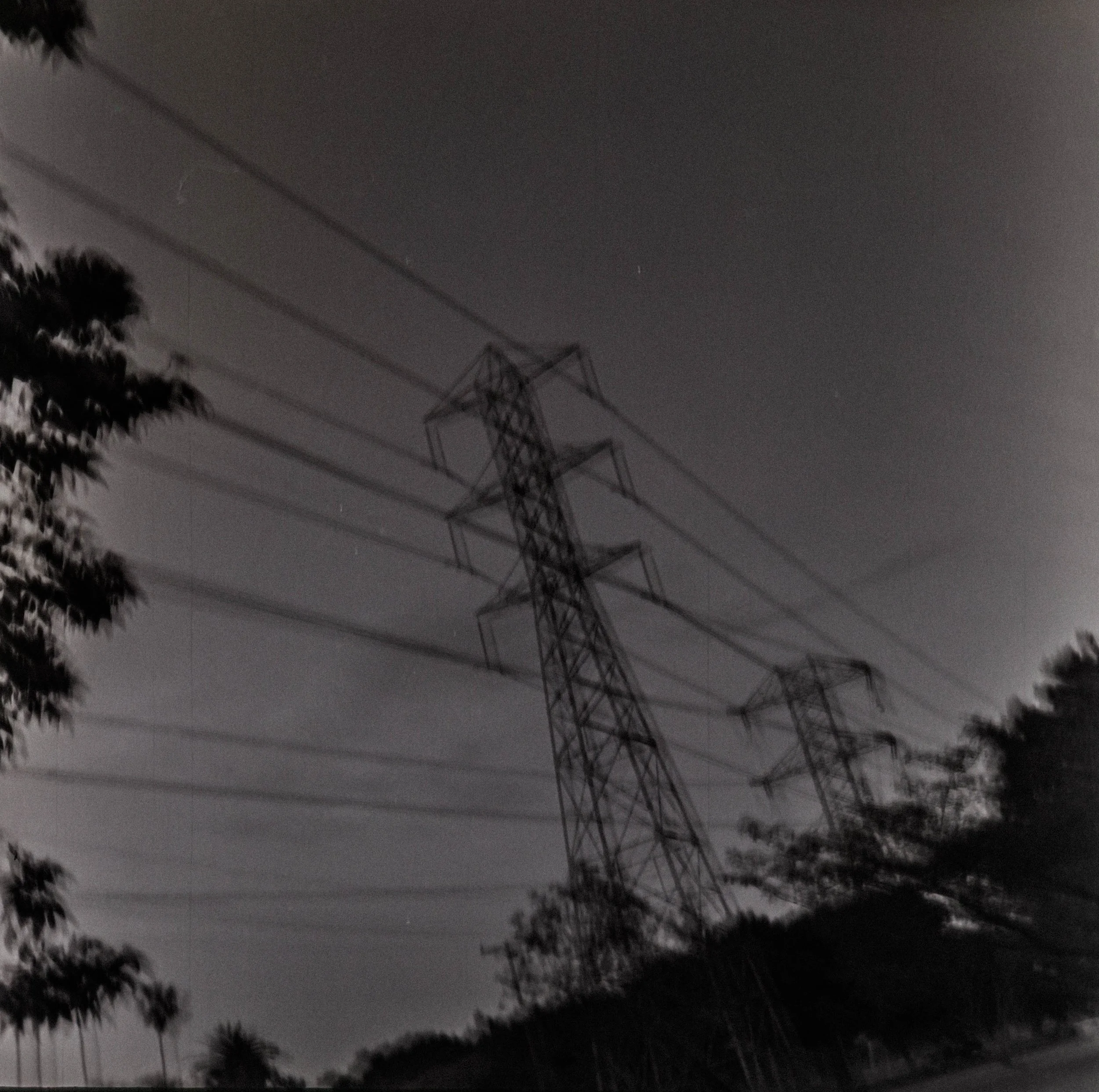 Nighttime black-and-white photo of electrical transmission towers and power lines among trees under a cloudy sky.