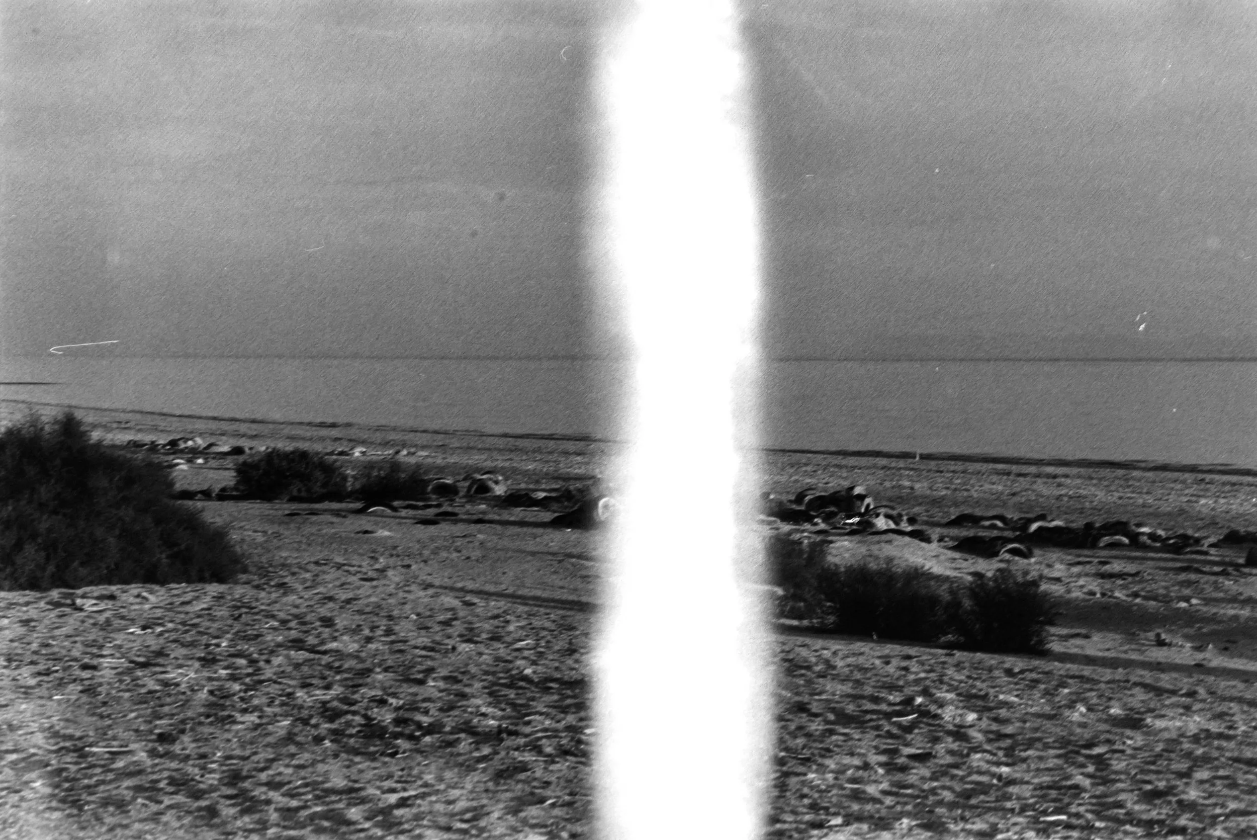 Black and white photograph of a beach with cars parked and dunes in the foreground, ocean and sky in the background, with a vertical white light streak down the middle.