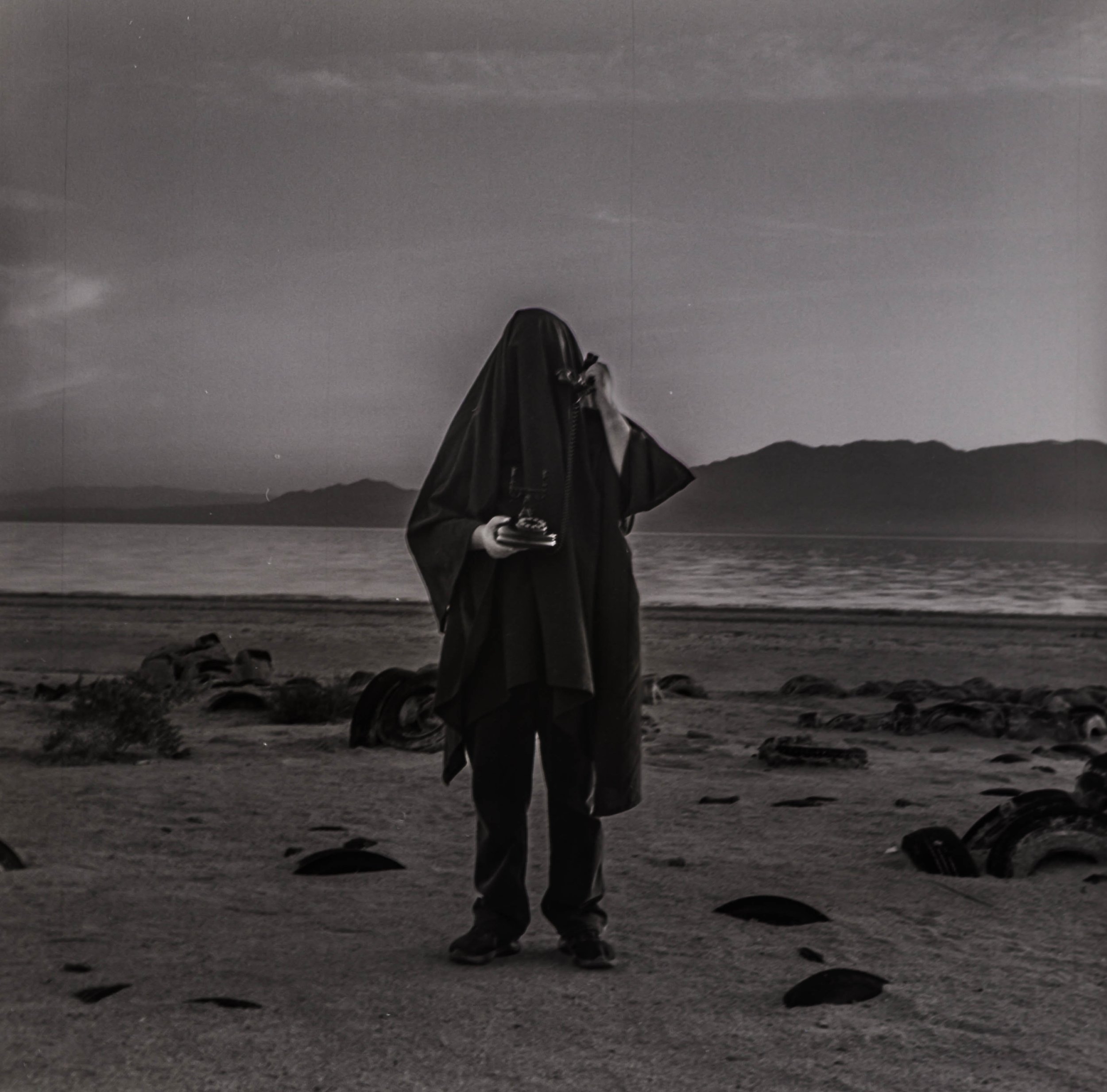 A person with a shrouded face holding a telephone on a deserted beach, with ruins scattered on the sand and mountains in the distance, in black and white.