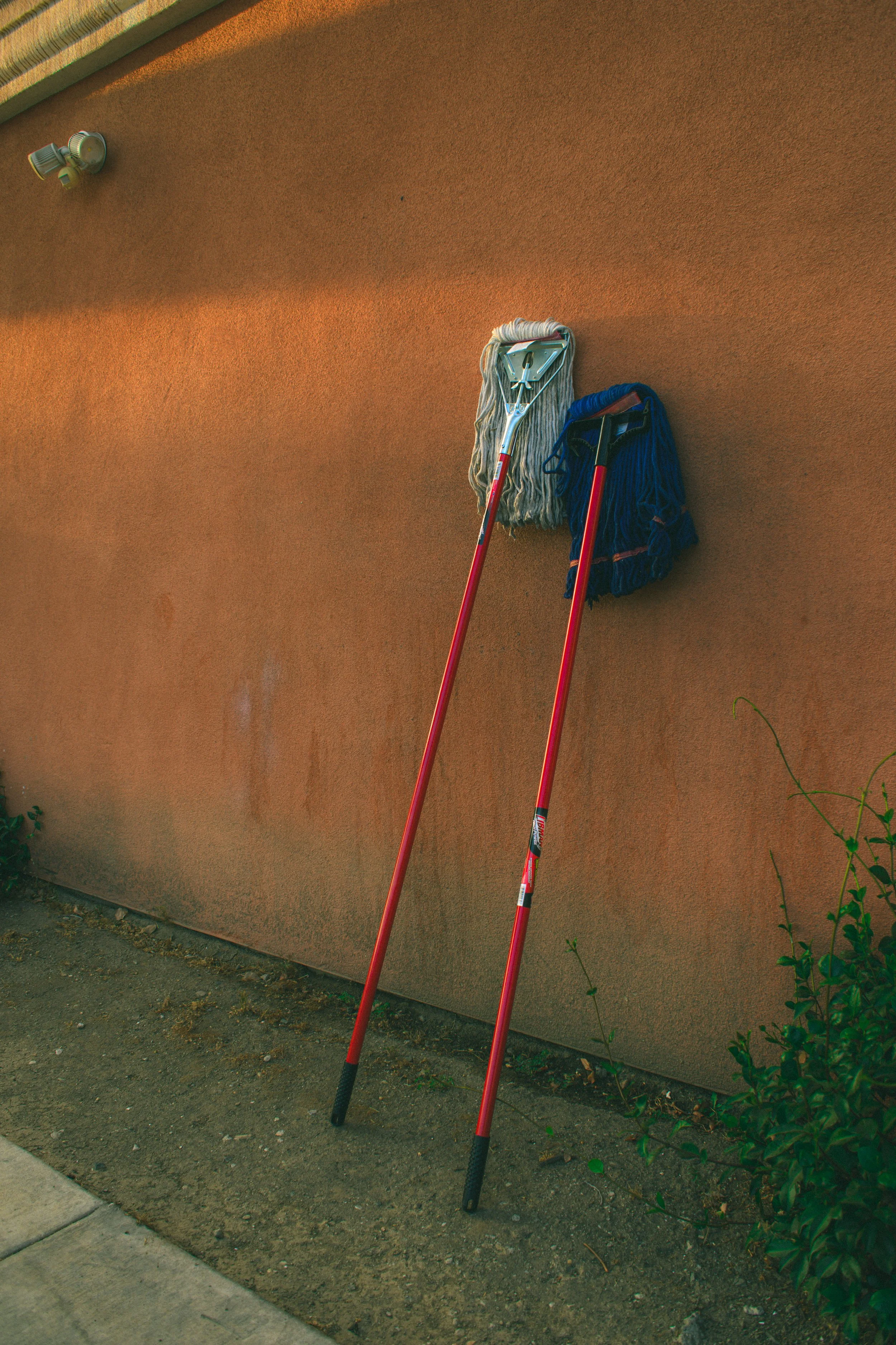 A pair of cleaning mops leaning against a textured orange wall outdoors, with a shrub and a concrete sidewalk in the foreground.