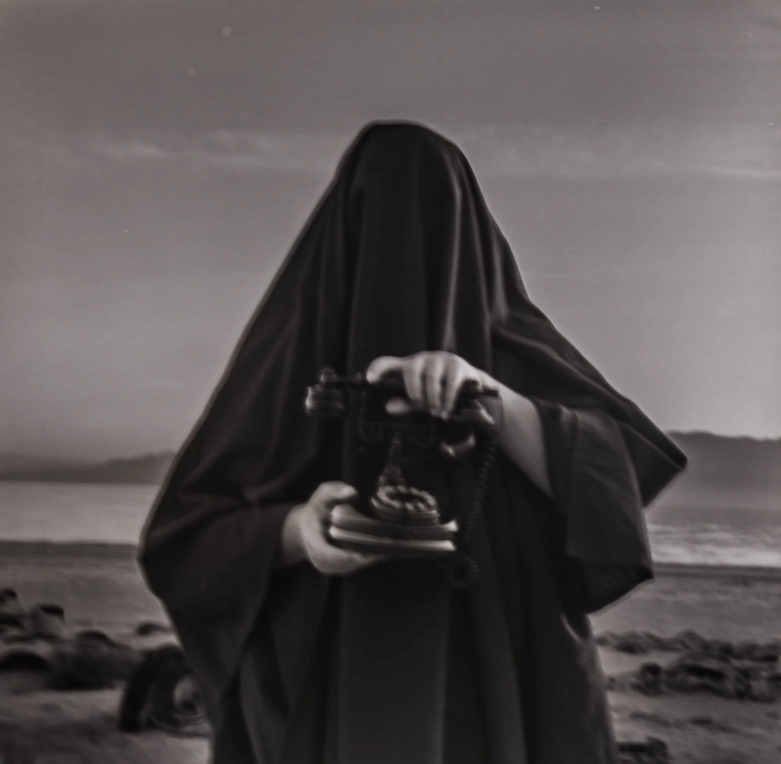 Person dressed as a ghost holding a vintage rotary phone, on a beach at sunset, black and white.