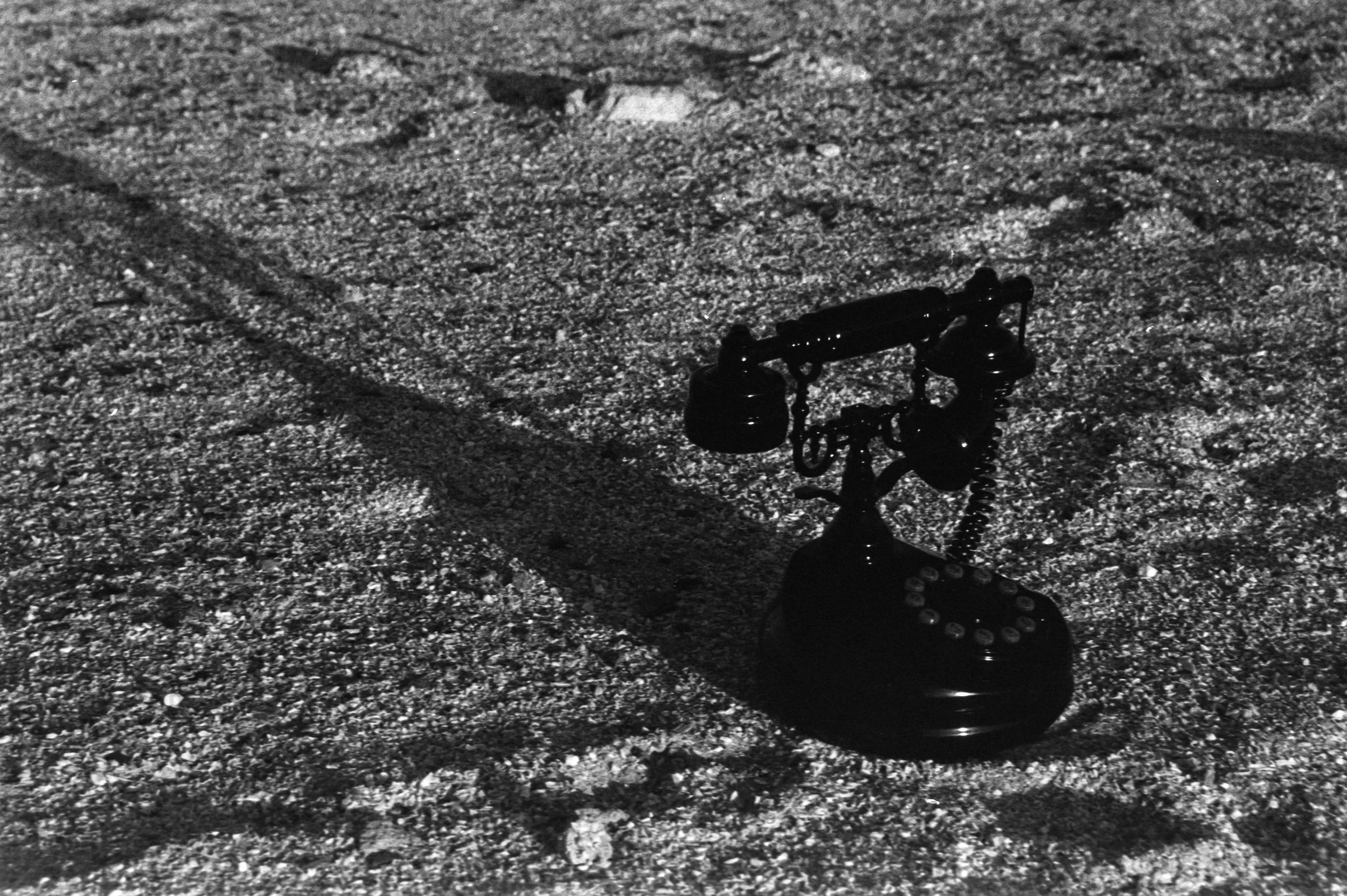 A shadow of a vintage telephone on a textured ground surface, photographed in black and white.
