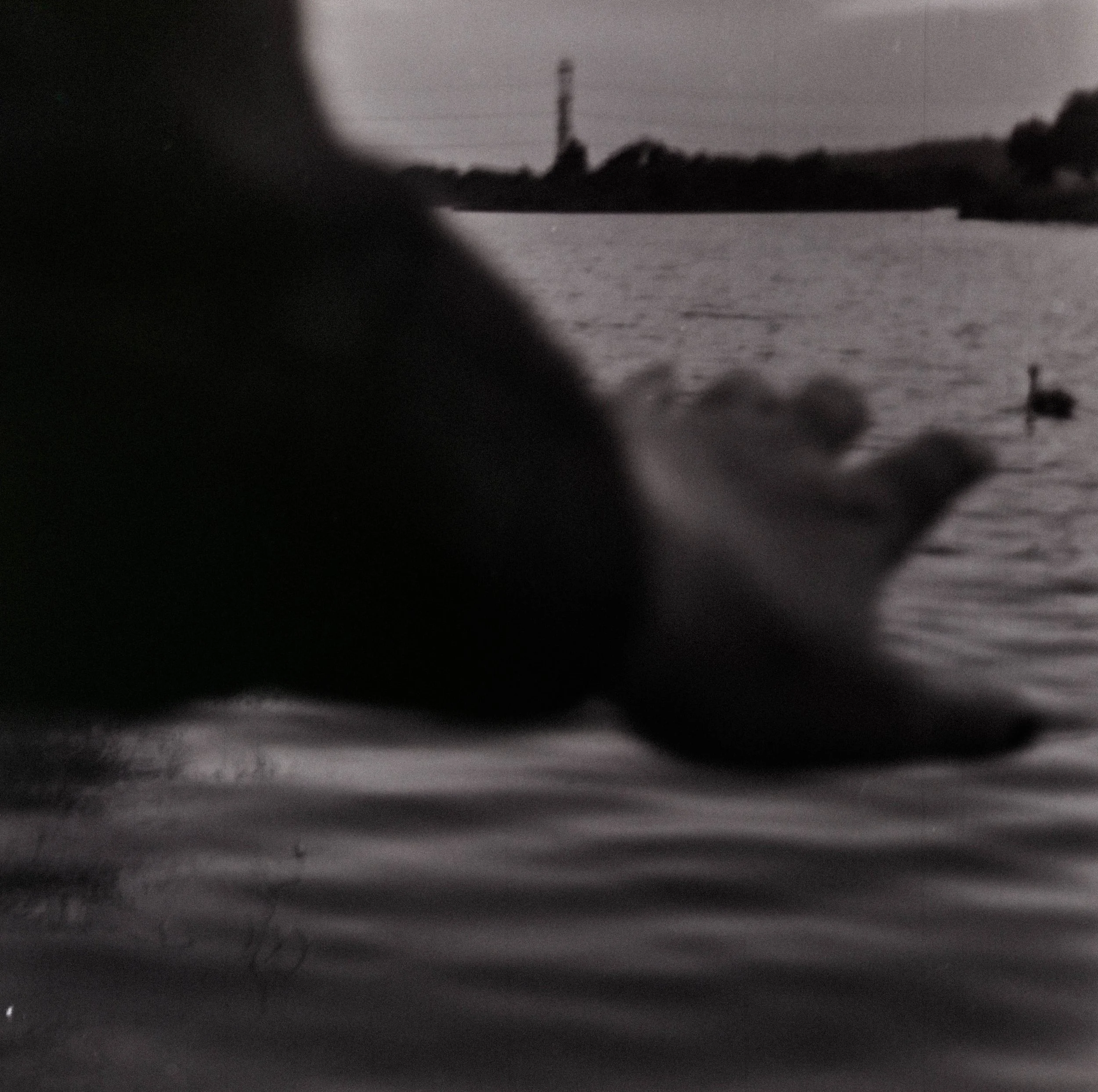 Close-up of a hand reaching into or resting on a body of water with a blurred background of a person on a boat and a power line tower in the distance.