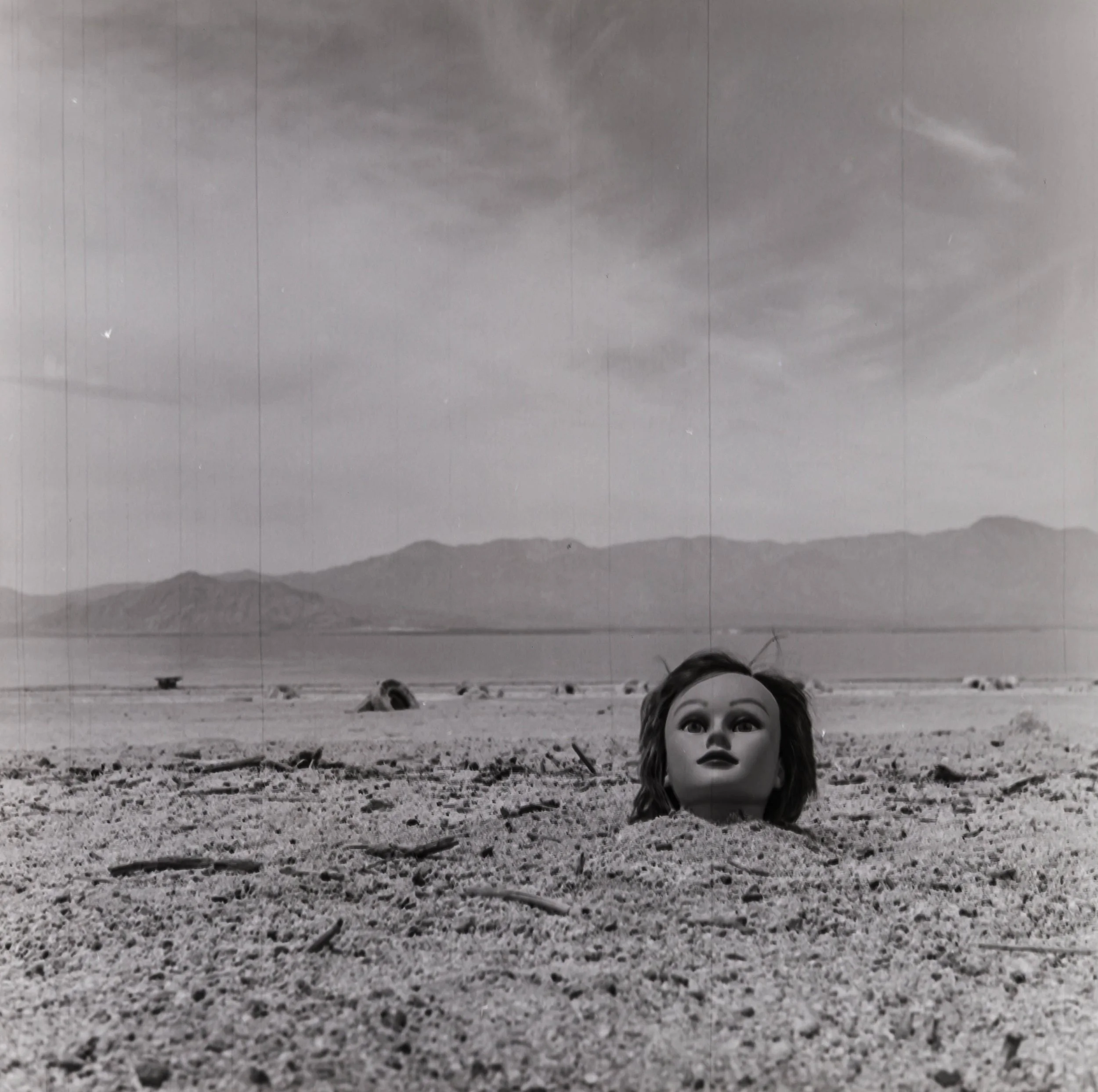 A doll's head partially buried in sandy ground with mountains and a cloudy sky in the background.