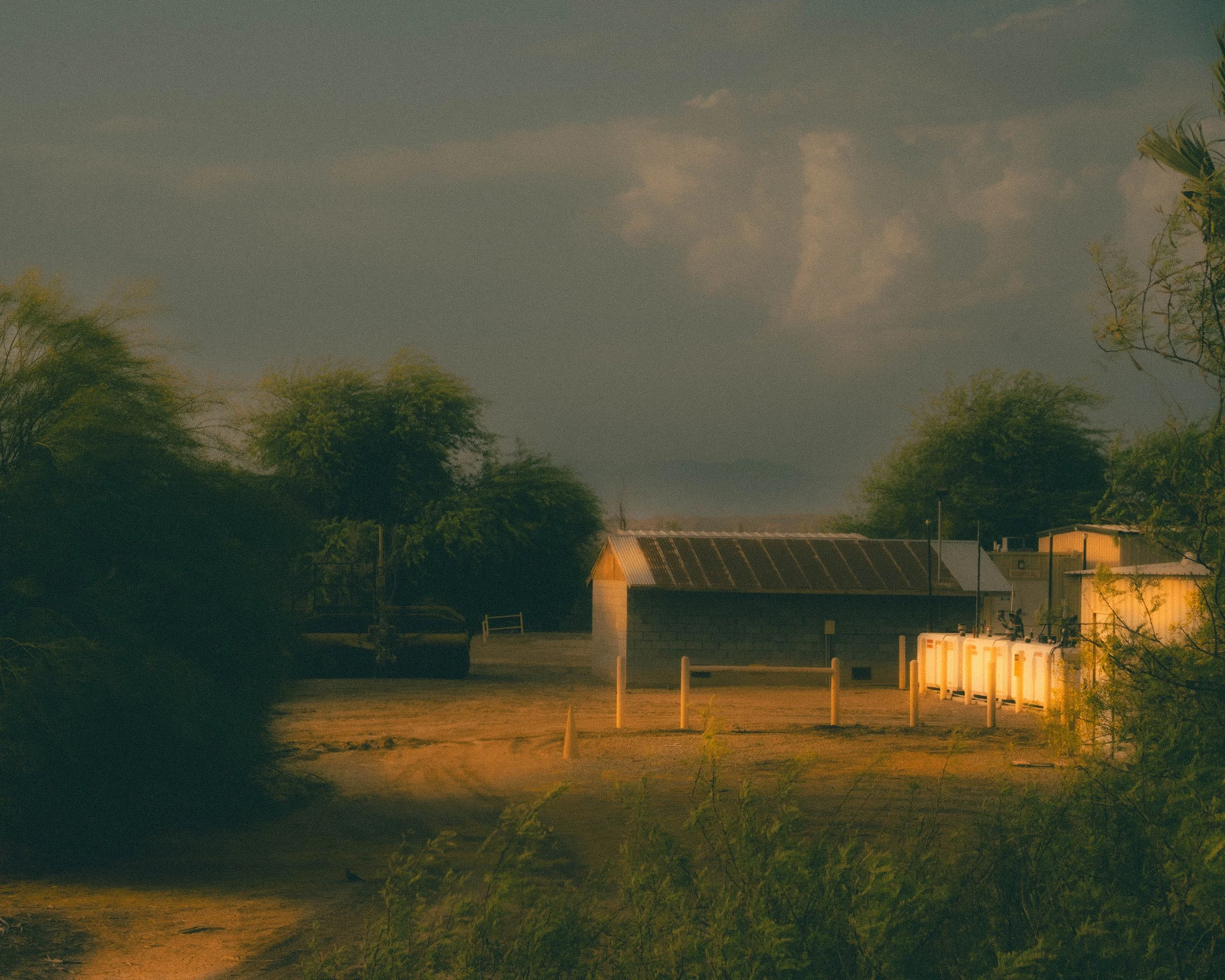 A rural scene with a dirt path leading to small buildings, trees, and a partly cloudy sky, lit by warm sunlight.