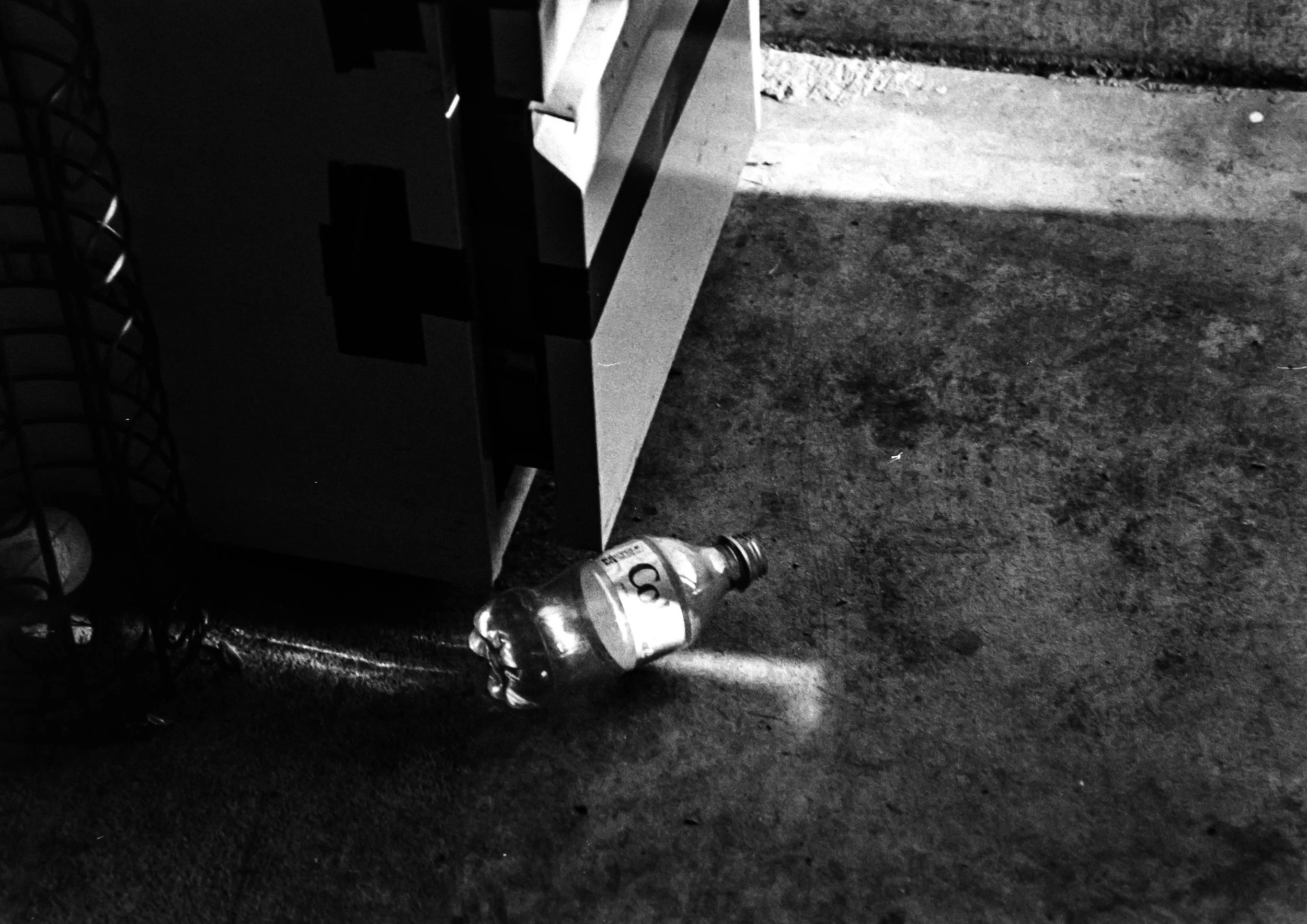 A discarded glass soda bottle lying on a dirty concrete floor near a piece of furniture and a wire basket.