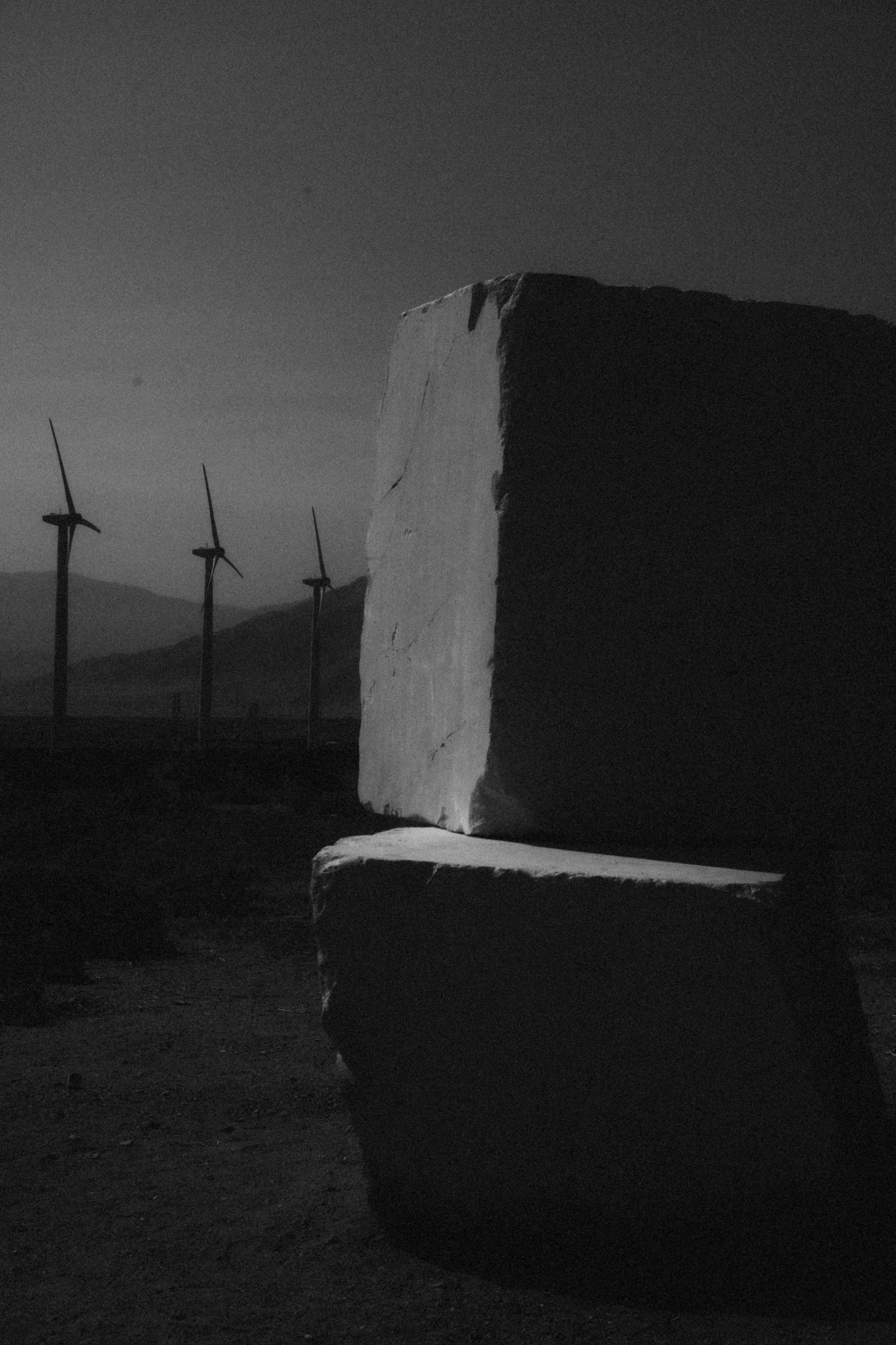 A large upright stone slab and a smaller slab beneath it in the foreground, with wind turbines and mountains in the distance under a clear sky.