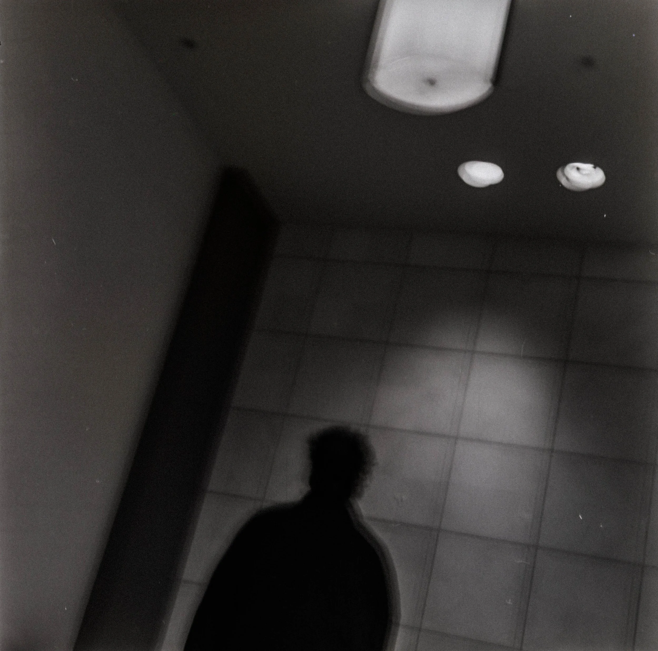 Silhouette of a person's head and shoulders reflected in a mirror on a tiled wall, with ceiling lights above.