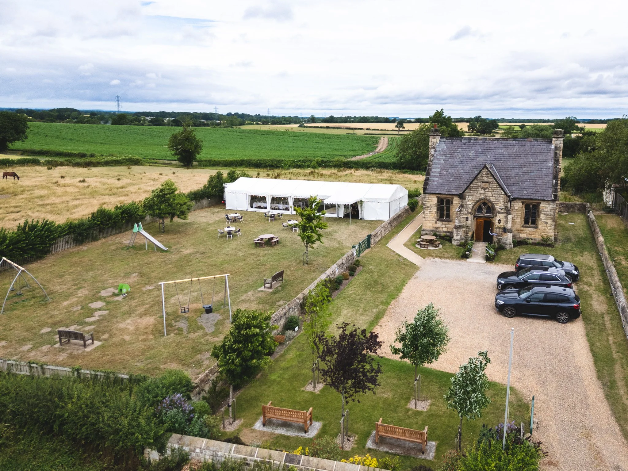 Aerial view of a countryside house with a gravel driveway, parked cars, a lawn with benches, trees, and a playground. A large white event tent is set up outside, surrounded by green fields and farmland.