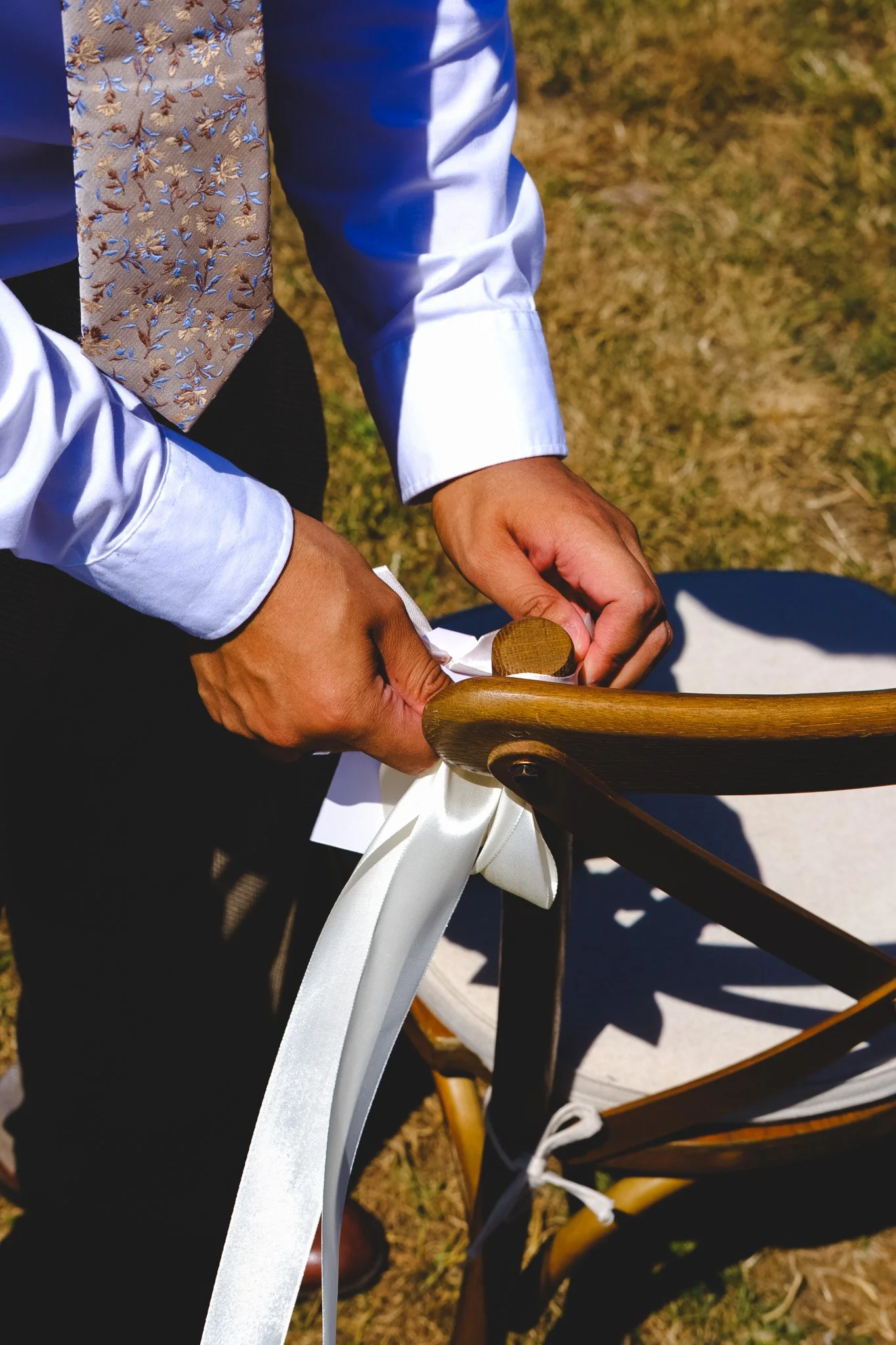 Person tying white satin ribbon on a wooden outdoor chair during a wedding ceremony, visible grass in background.
