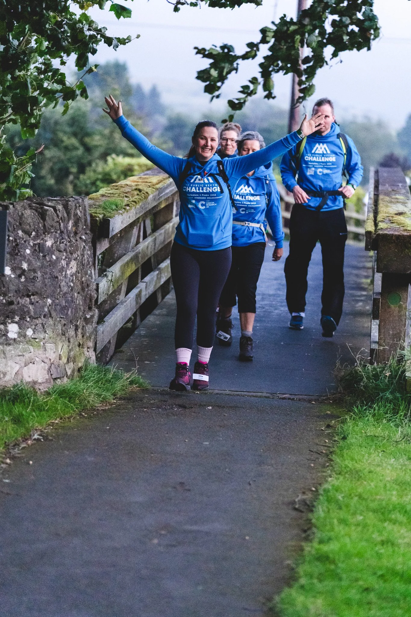 A group of four people walking outdoors on a narrow path, wearing matching blue jackets with a logo, smiling and waving as they walk.