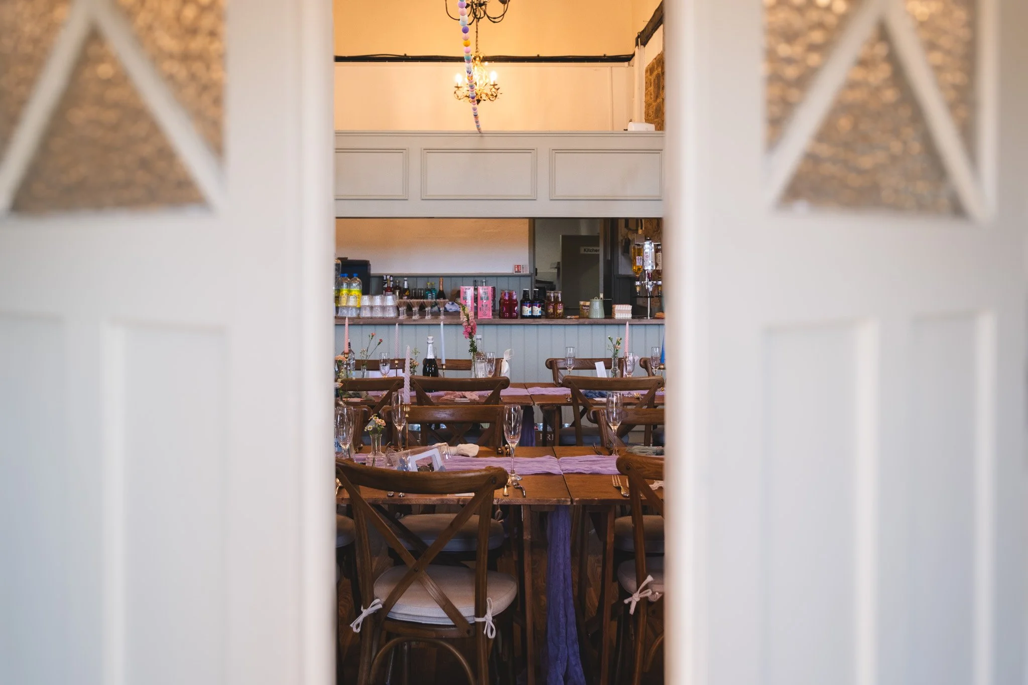A dining area viewed through a partially open door, with tables set with glassware, flowers, and pink tablecloths, and a bar in the background.