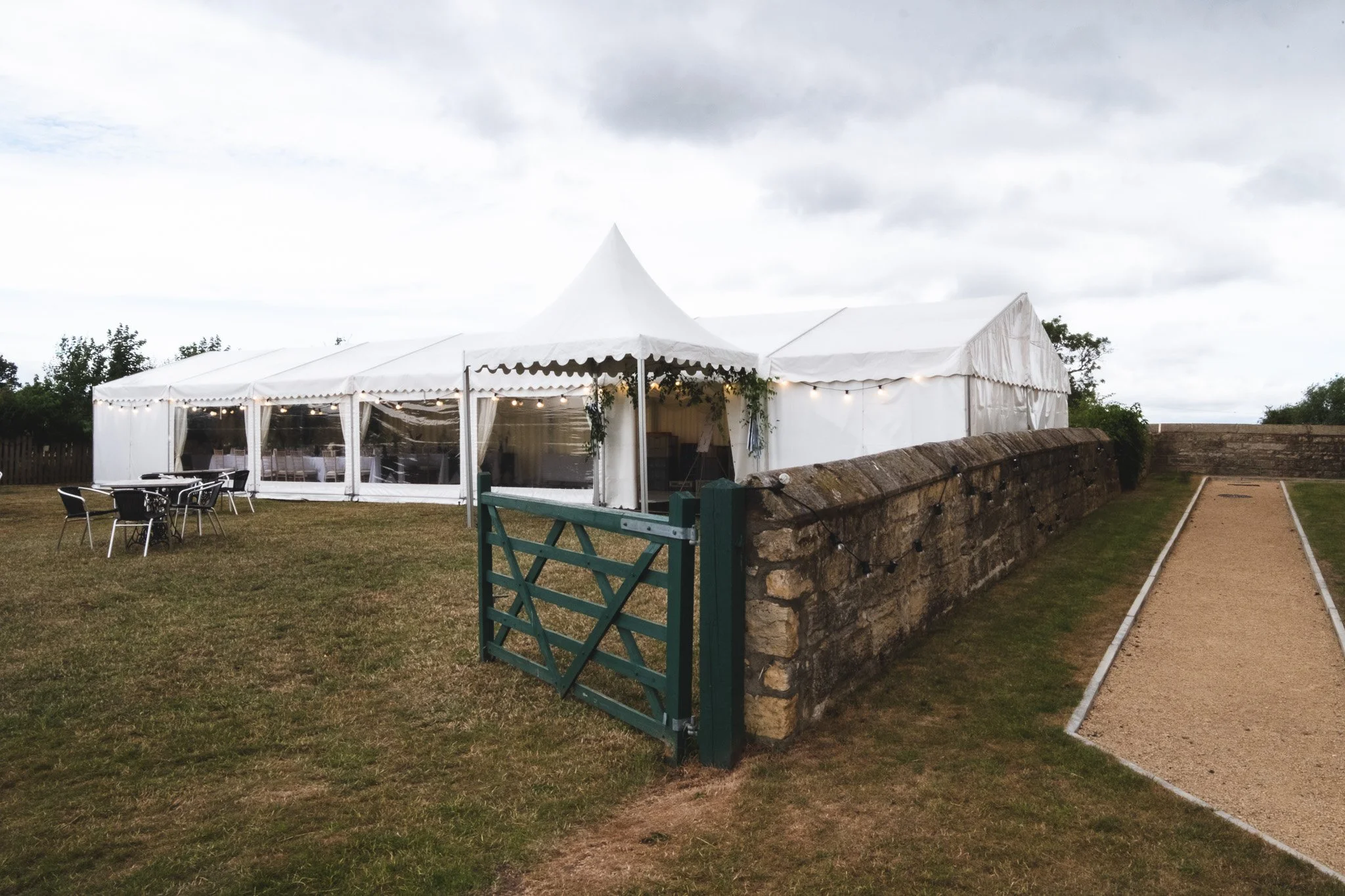 White event tent with string lights set up on a grassy lawn, with outdoor tables and chairs nearby and a stone wall on the right side under cloudy skies.
