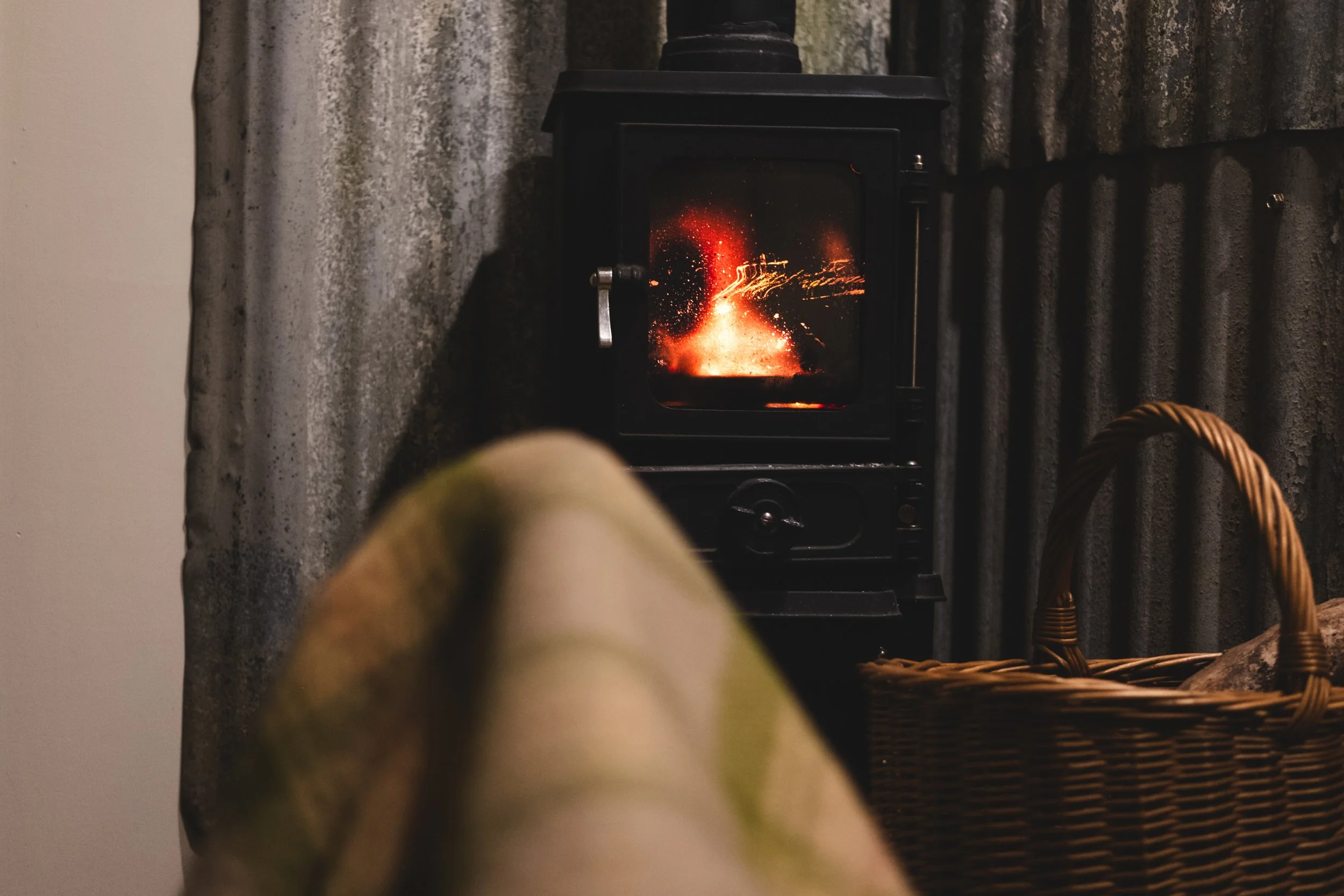 A small black wood stove with a glowing fire inside, situated next to a wicker basket on the right, with a beige cushioned chair or sofa in the foreground, and corrugated metal walls in the background.