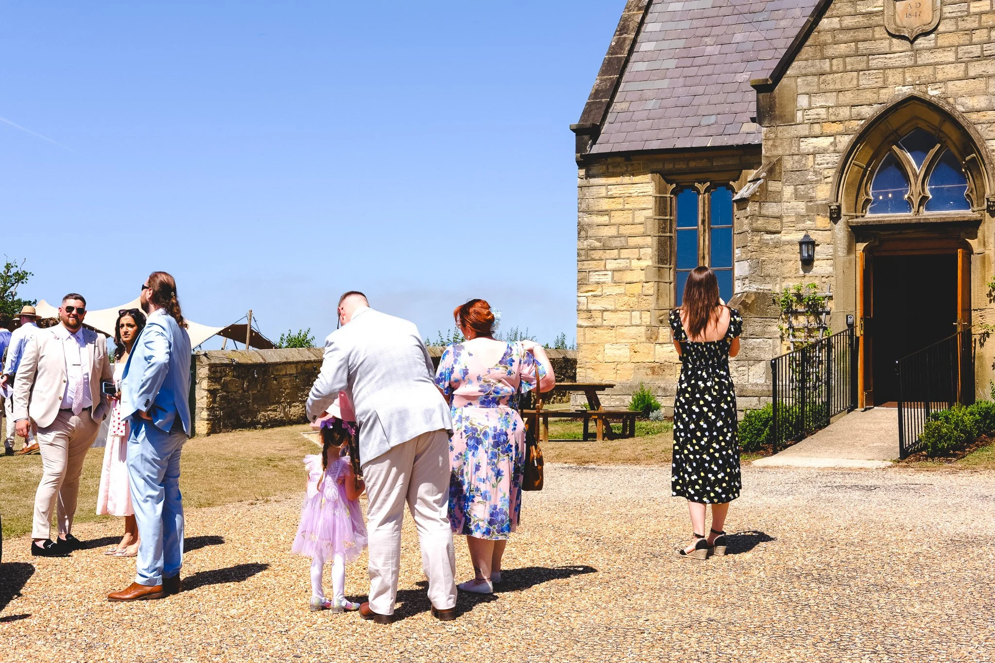 A group of people dressed in formal attire outside a stone church on a sunny day with clear blue sky.