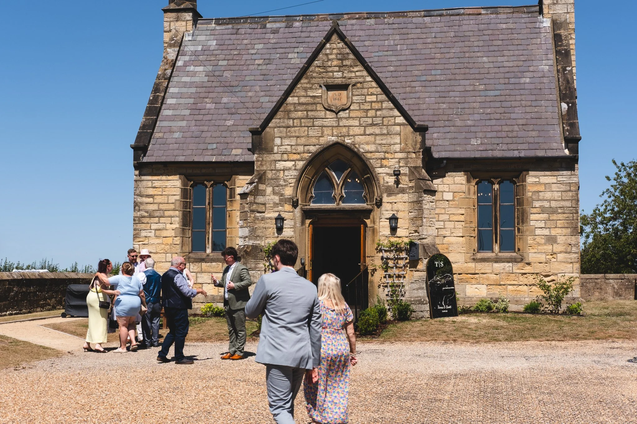 Group of people dressed in semi-formal attire gathering outside a stone church with gothic windows and a slate roof, on a sunny day.