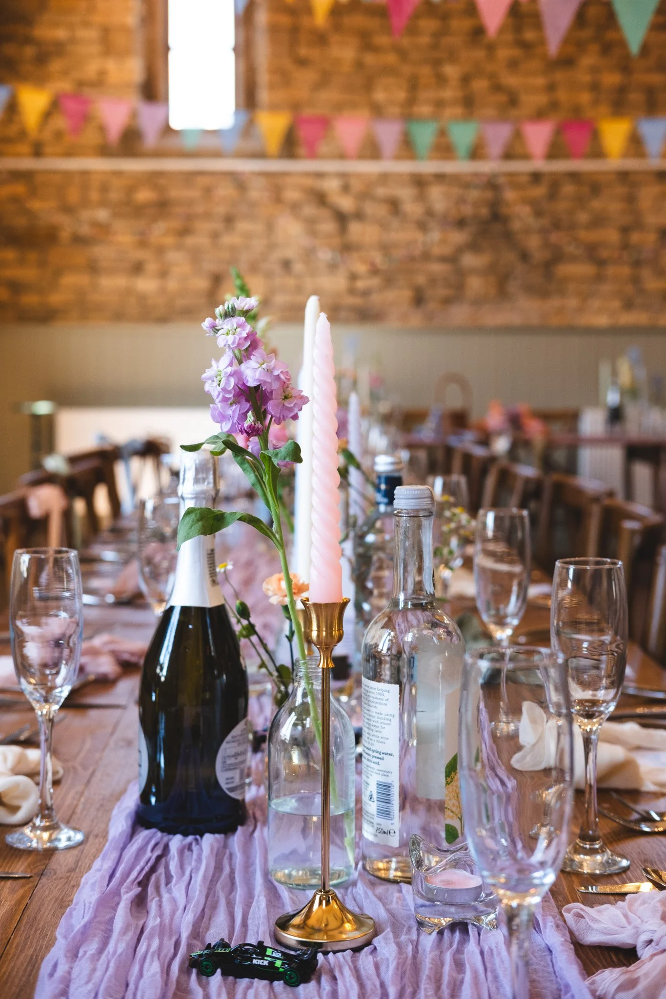 Table set for a celebration with pink and purple flowers, candles, glasses, bottles, and a purple table runner in a rustic venue with string bunting.
