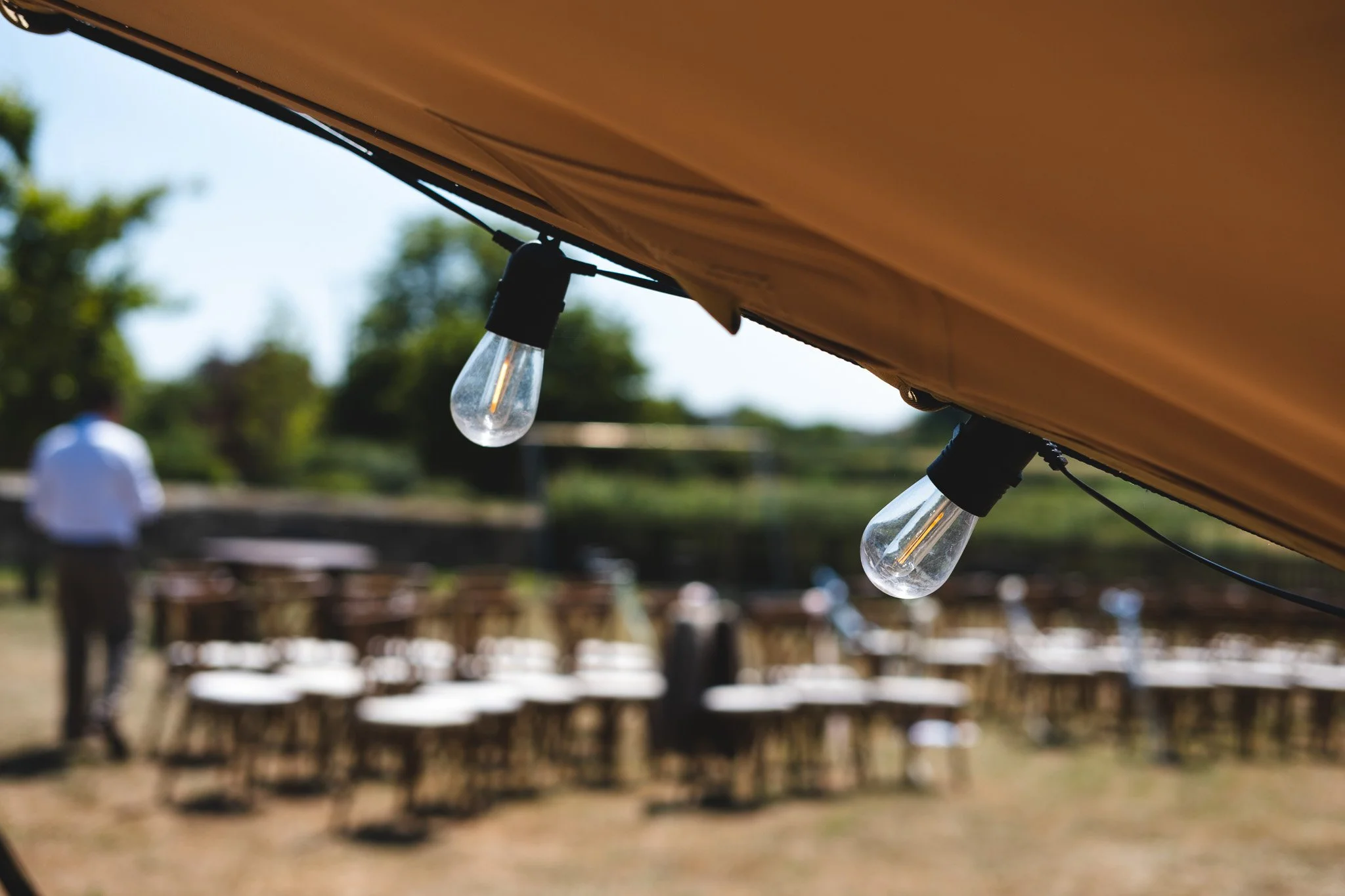 String of outdoor string lights hanging under a canopy with blurred chairs and a person in the background.