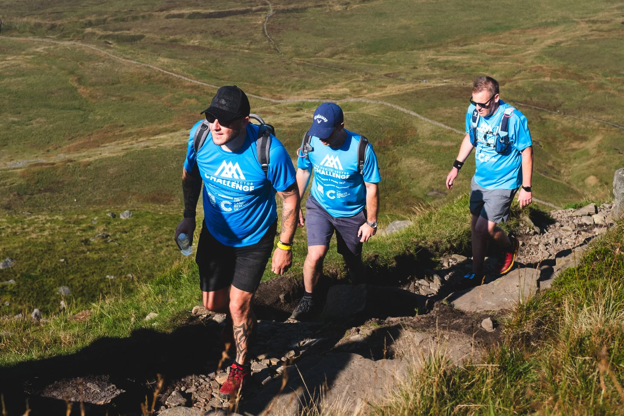 Three men wearing blue event shirts hiking on a rocky trail in a green hillside landscape.