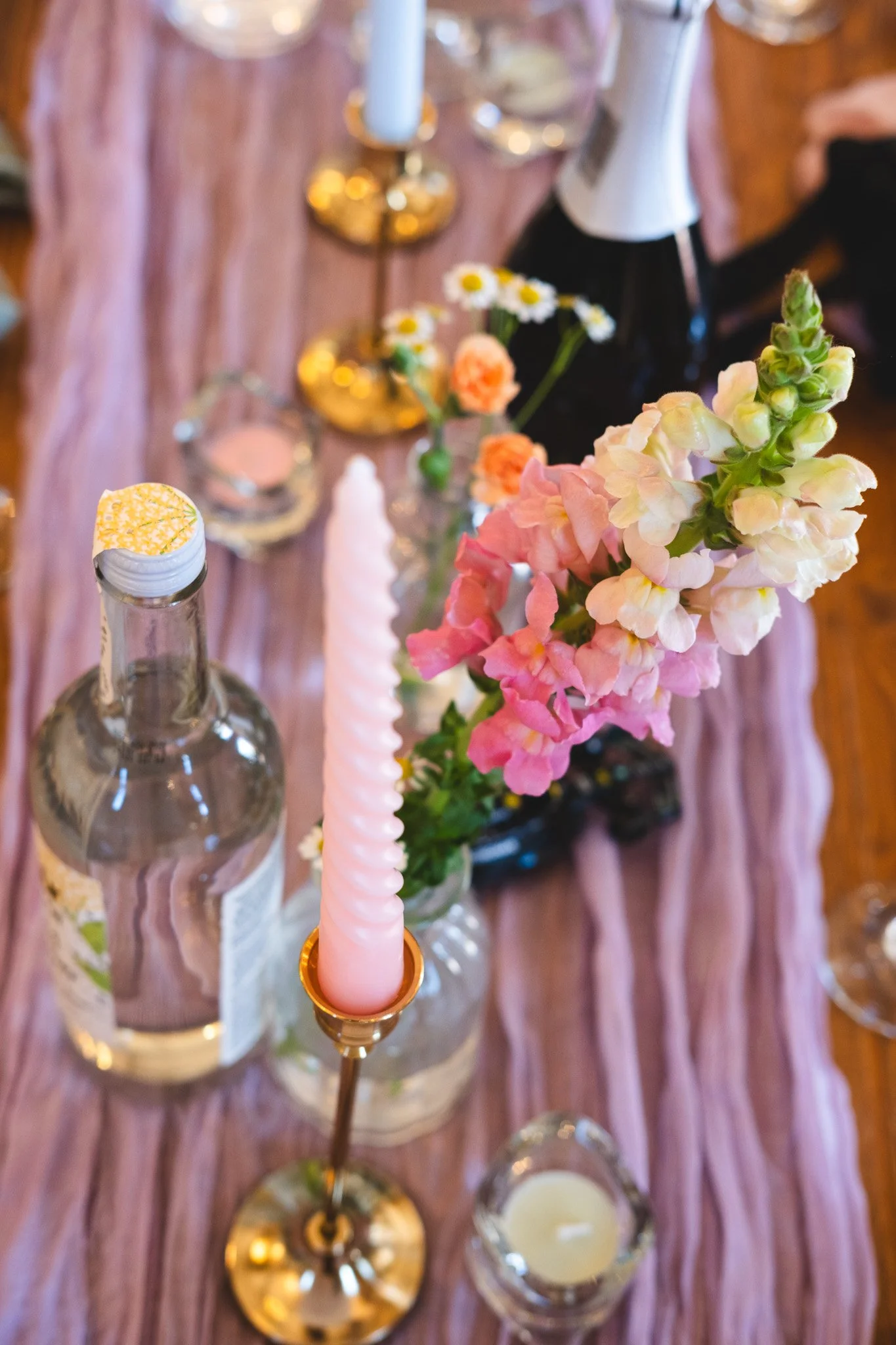 A table decorated with flowers, candles, and bottles, featuring a pink twisted candle in a gold holder, pink flowers, and small vases on a pink table runner.