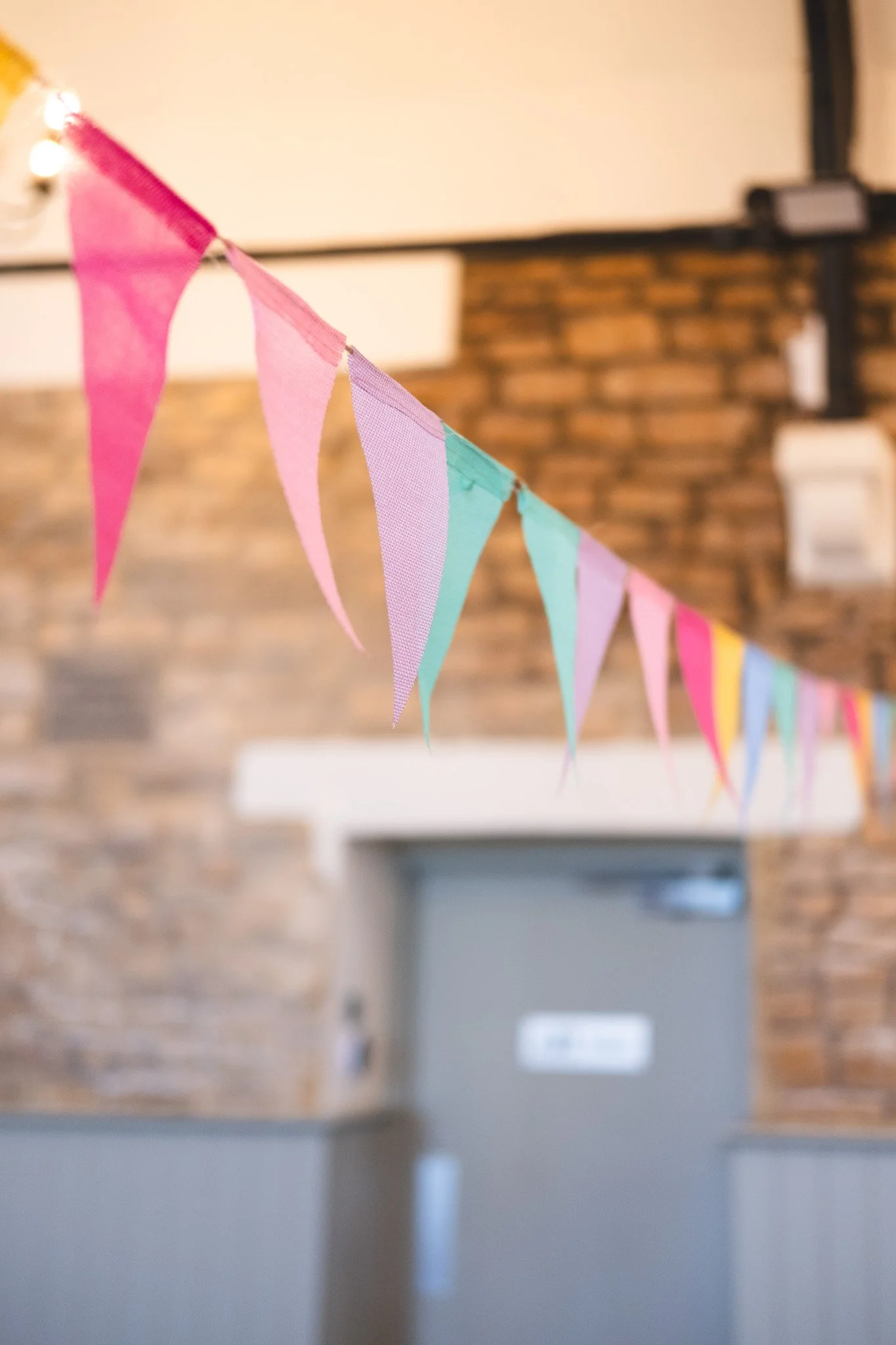 Colorful fabric pennant banner hanging in front of a brick wall, with a blurry background of a room interior.