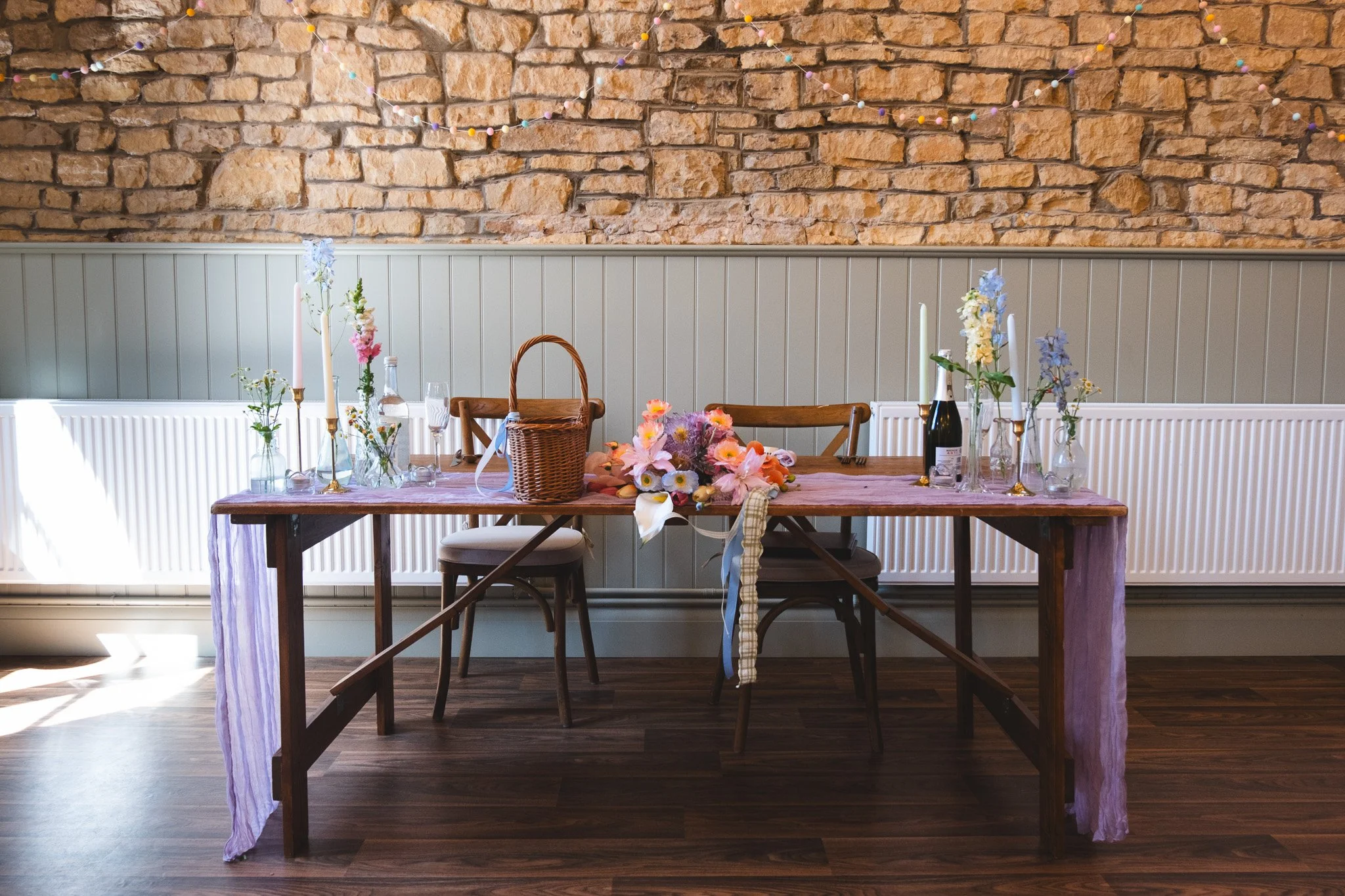A decorated wooden dining table with floral arrangements and candles, set against a brick wall with string lights