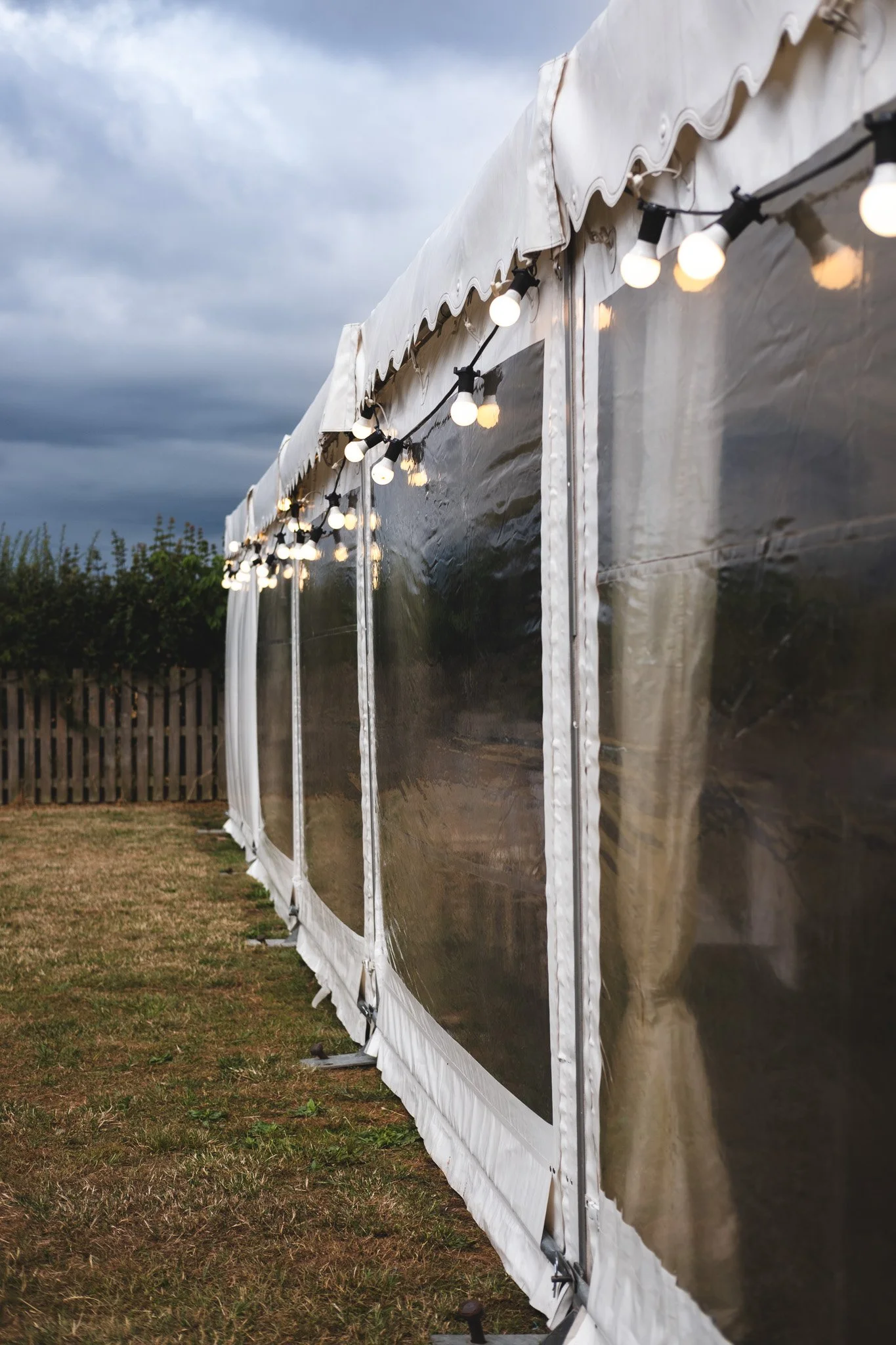 An outdoor tent with transparent plastic walls and a white canopy, decorated with string lights, on a grassy area with a cloudy sky overhead.