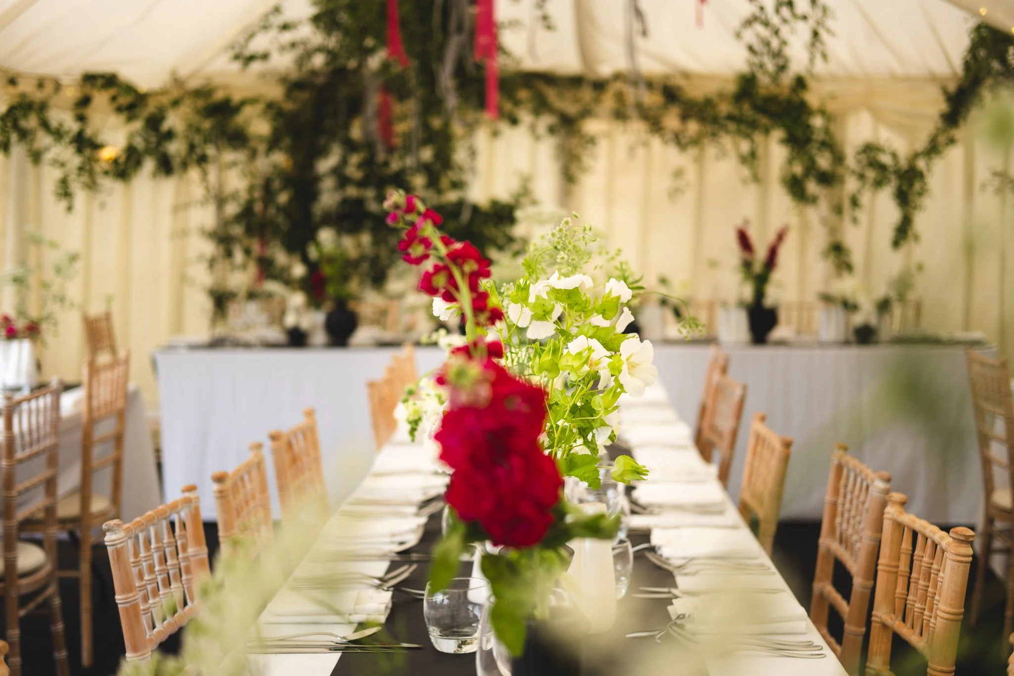 A decorated banquet table with floral centerpieces featuring red and white flowers, set with plates, silverware, and napkins in a decorated event tent.