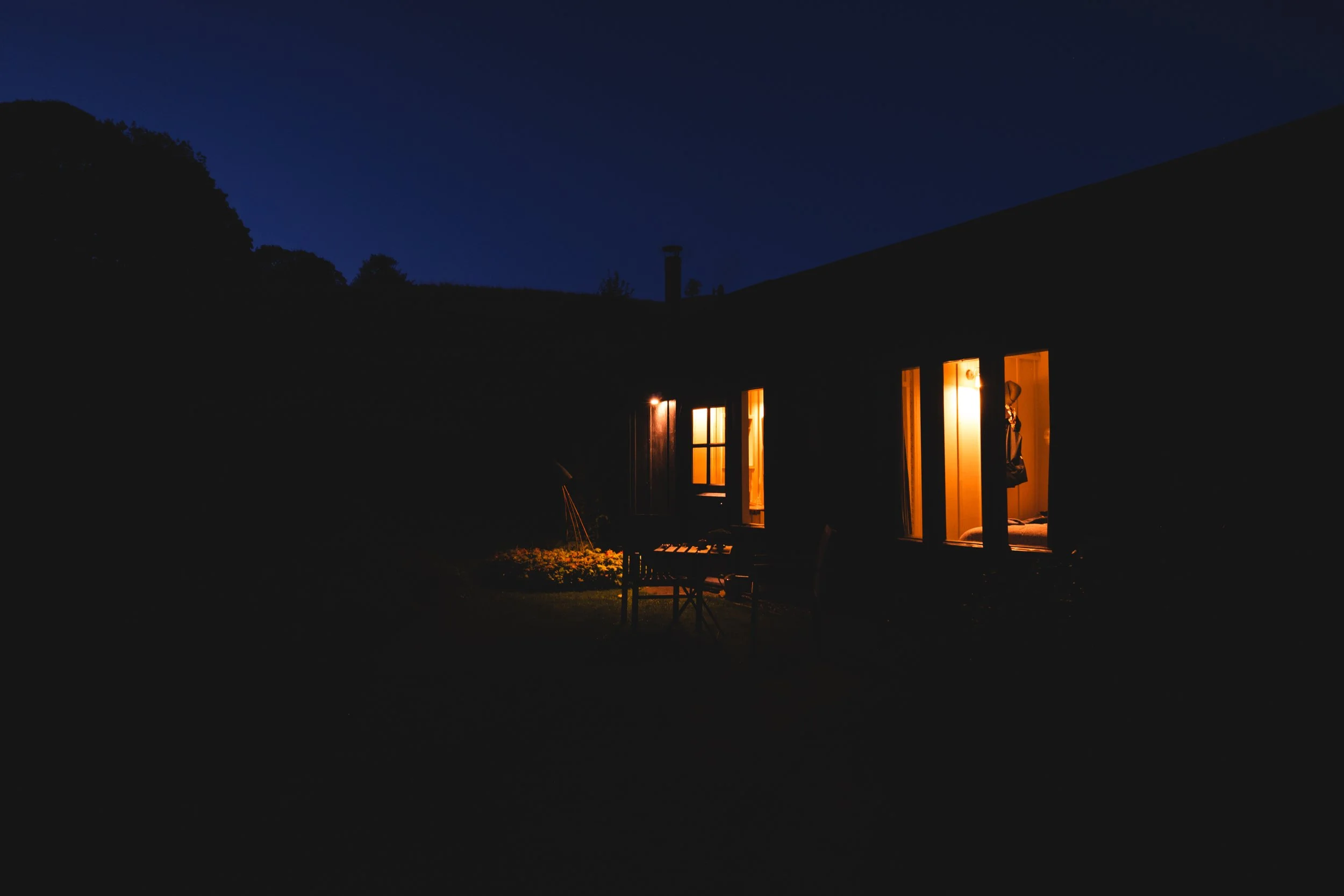Nighttime view of a house with three lit windows, warm yellow glow inside, dark surroundings, silhouette of trees and hills in the background.