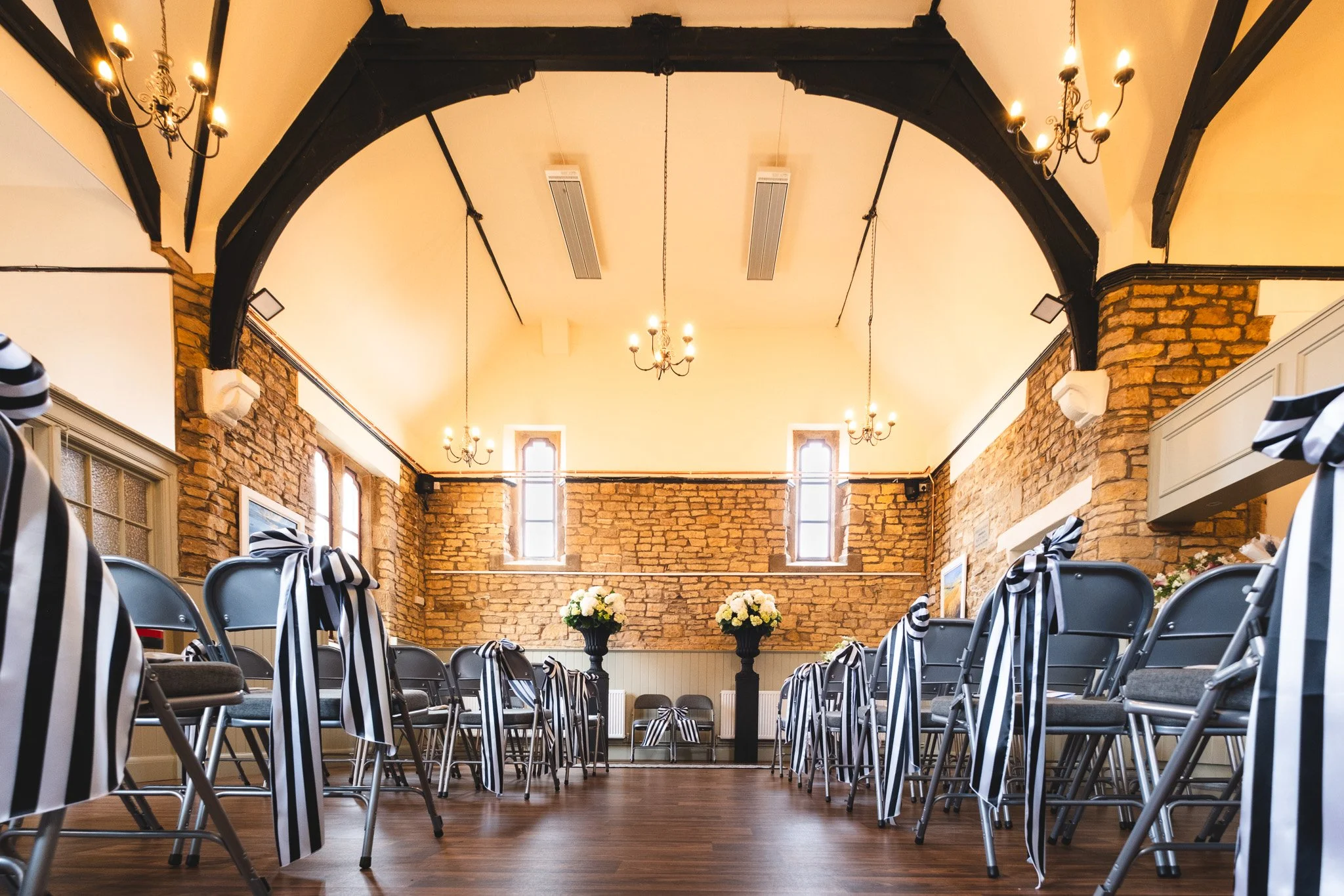 A decorated wedding or event ceremony space with rows of chairs tied with black and white striped ribbons, floral arrangements on pedestals, and a brick wall backdrop with tall windows. Chandeliers hang from the ceiling and the wood floor is visible.